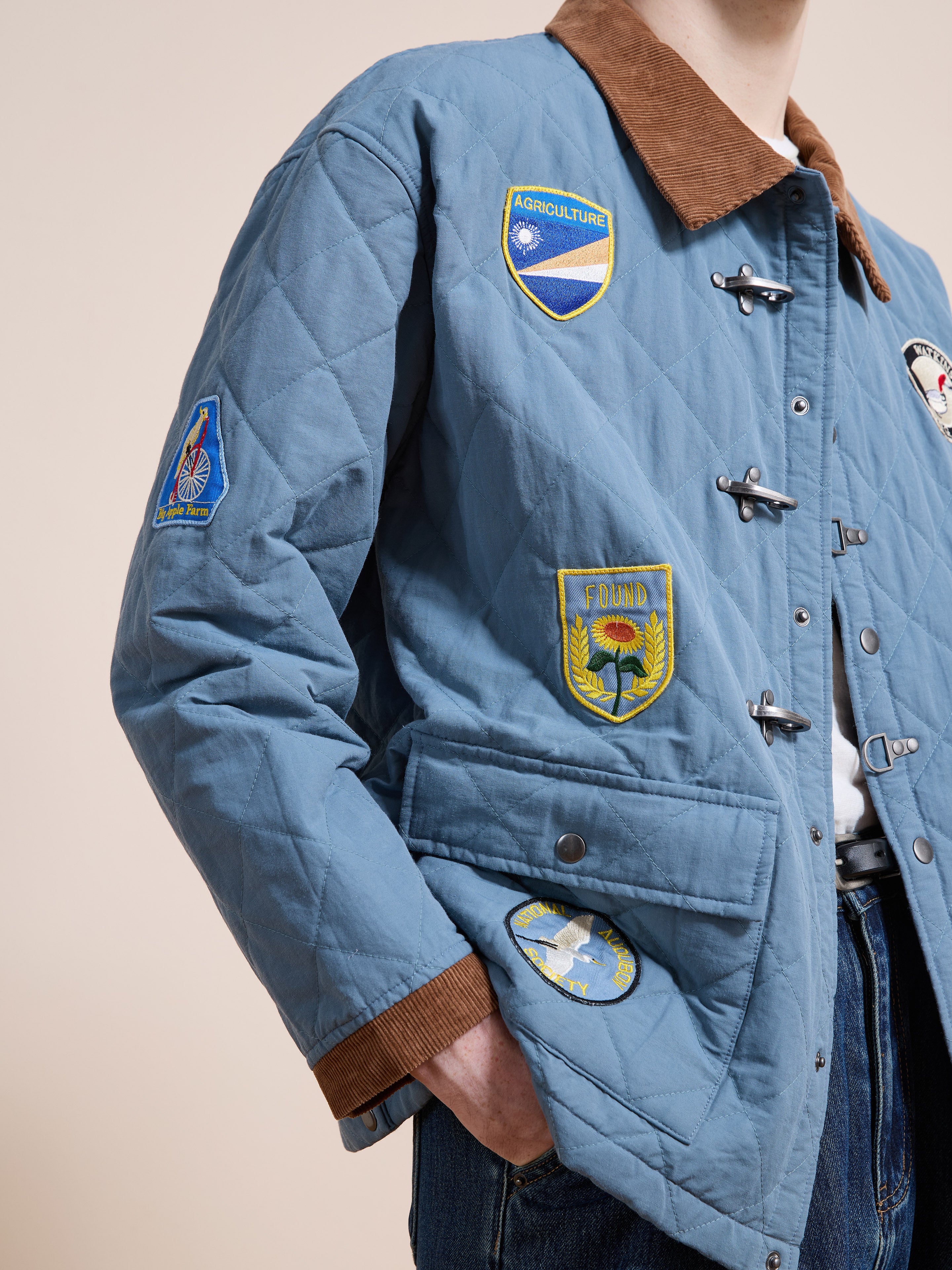 A person wears the FOUND Farmstead Quilt Patch Jacket—a blue quilted jacket with a brown collar and assorted embroidered patches—paired with jeans. Only the upper body is visible.