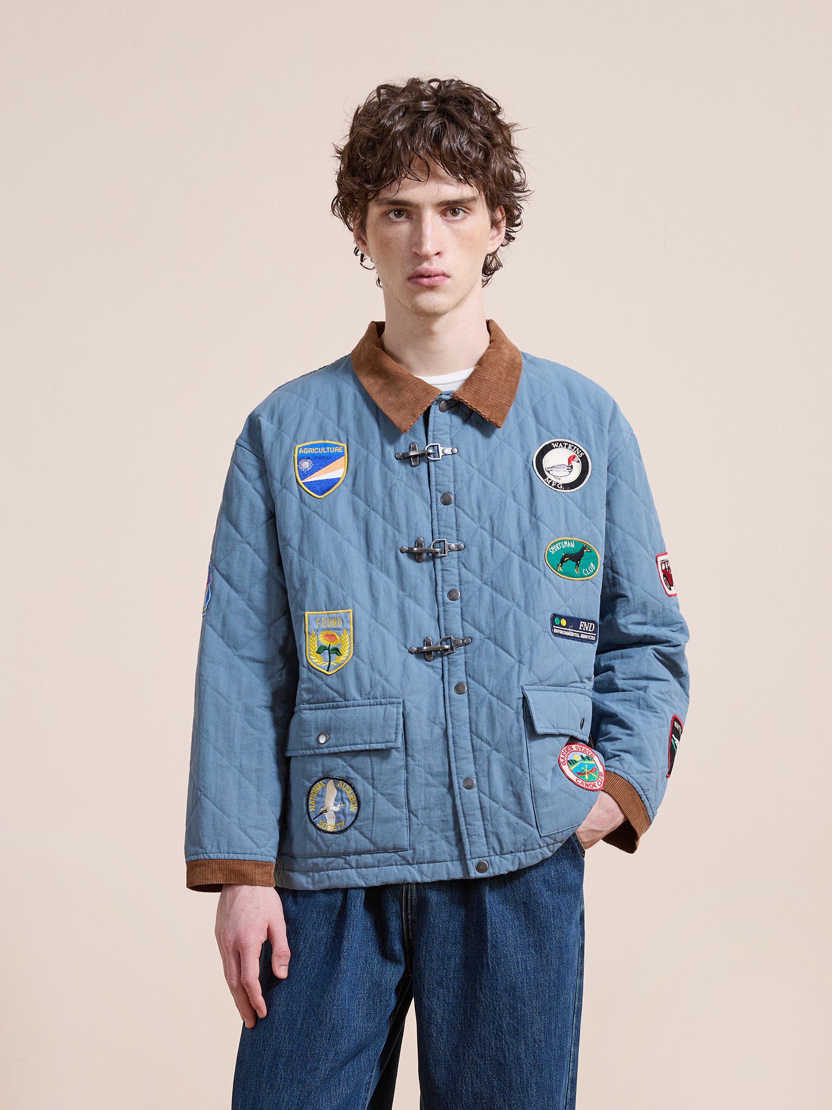 A young man wears the FOUND Farmstead Quilt Patch Jacket, featuring a light blue quilted design with colorful patches, brown collar and cuffs, and toggle buttons, standing against a plain beige background.