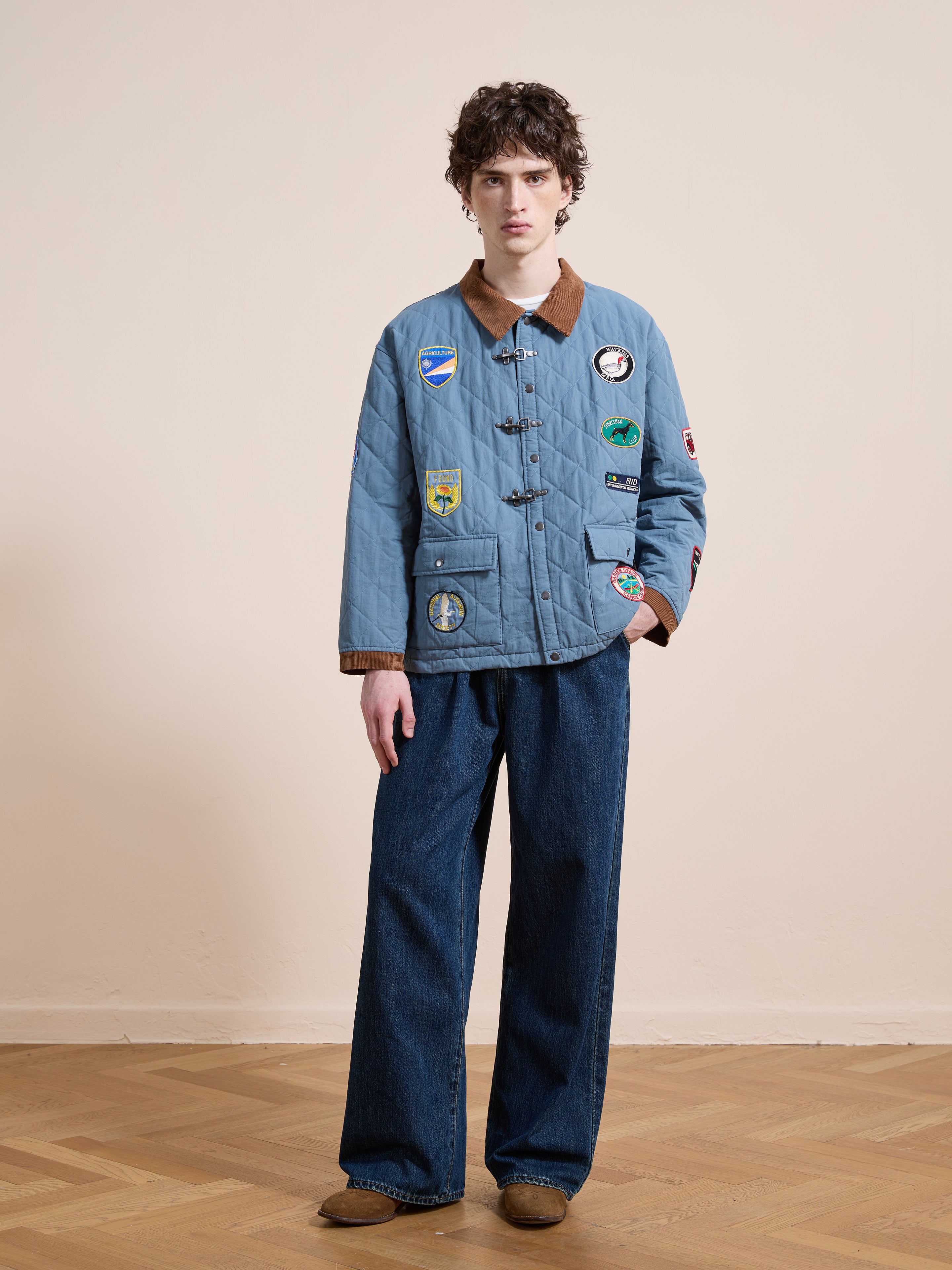 A person stands indoors against a beige wall and wooden floor, wearing the FOUND Farmstead Quilt Patch Jacket with various patches, wide-leg blue jeans, and brown shoes.