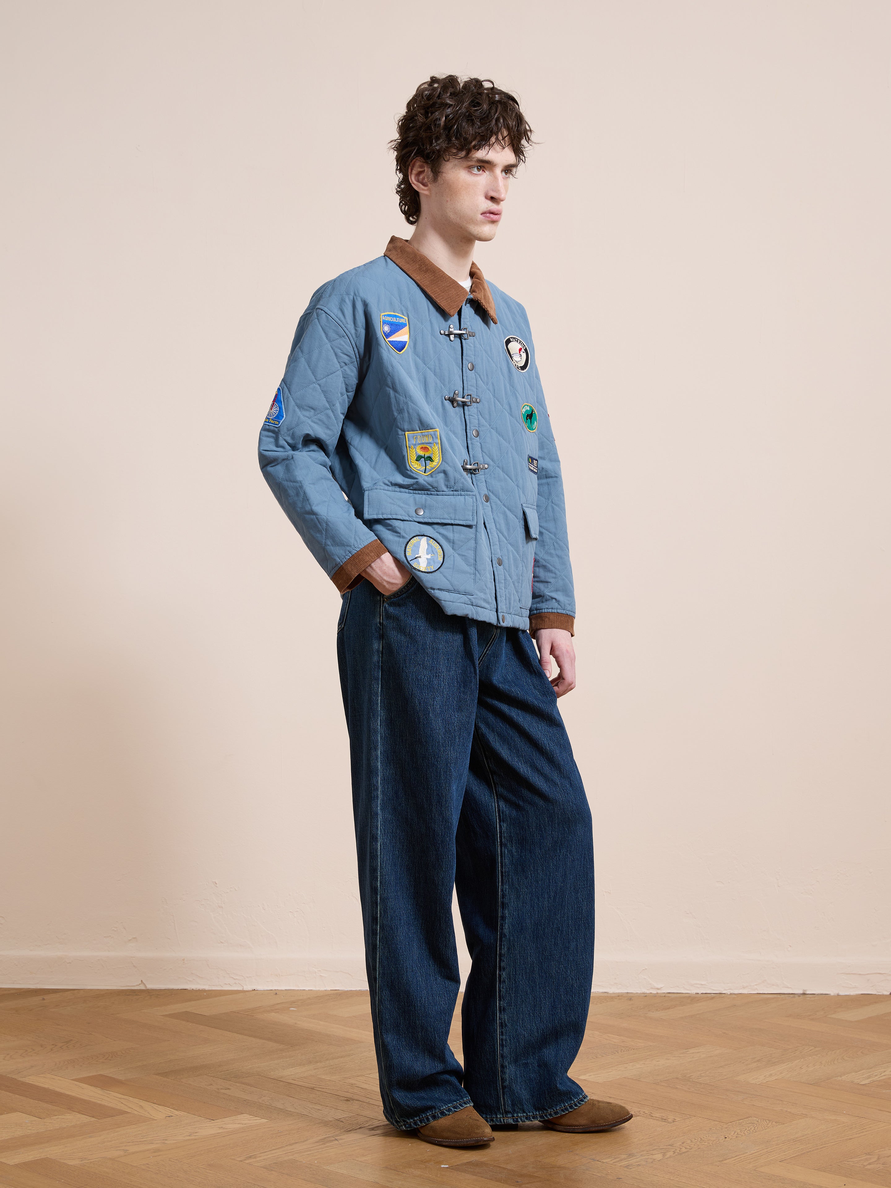 A man indoors wears the FOUND Farmstead Quilt Patch Jacket, styled with wide-leg blue jeans and brown shoes, standing on a wooden floor against a plain beige wall.