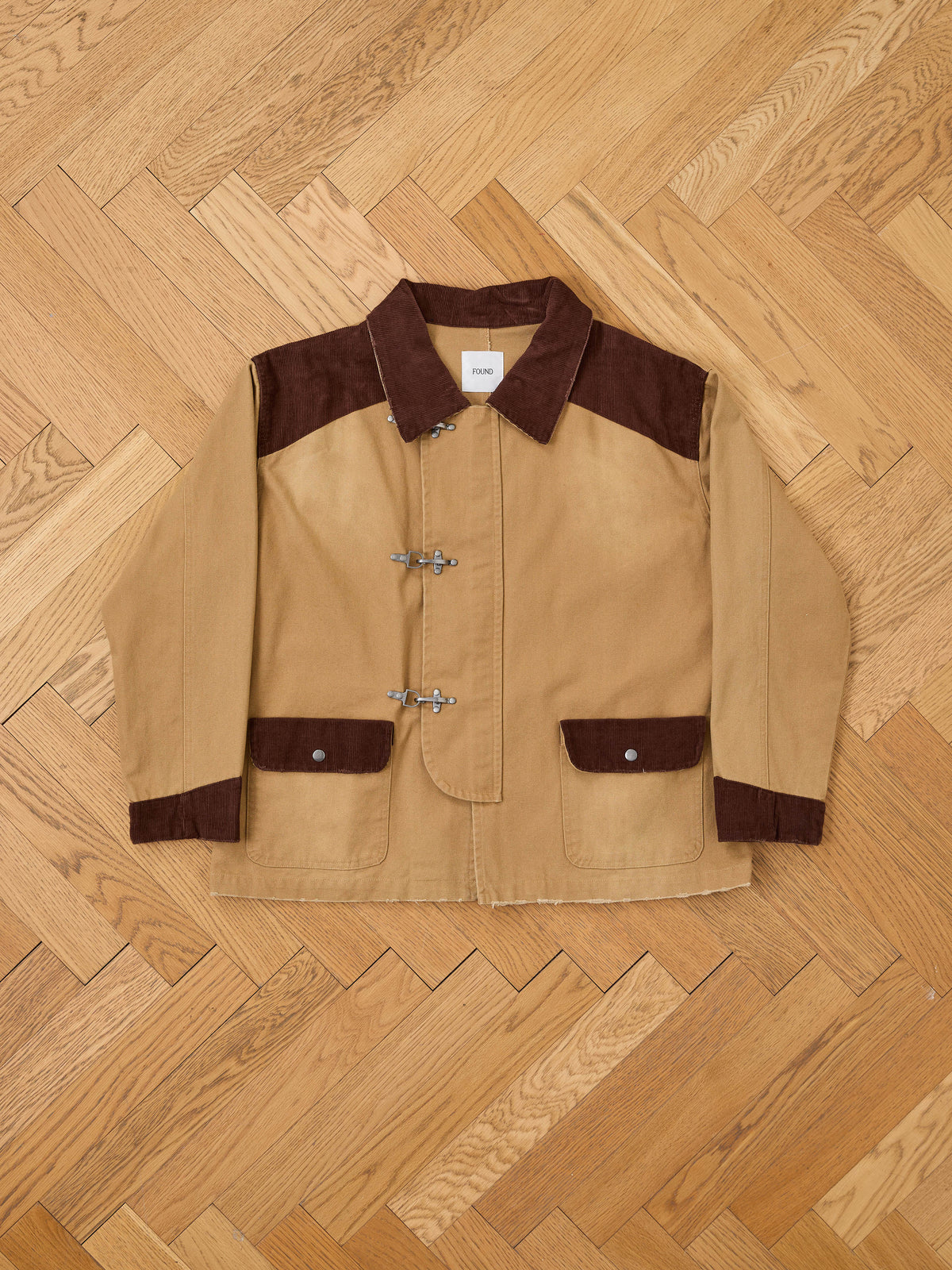 The Fireman Clasp Canvas Work Jacket by Found features a tan washed canvas body with contrasting dark brown collar, shoulders, and pockets. Designed with three signature metal fireman clasp closures, this mid-weight jacket offers a unisex relaxed fit and is displayed laid flat on a wood herringbone floor.