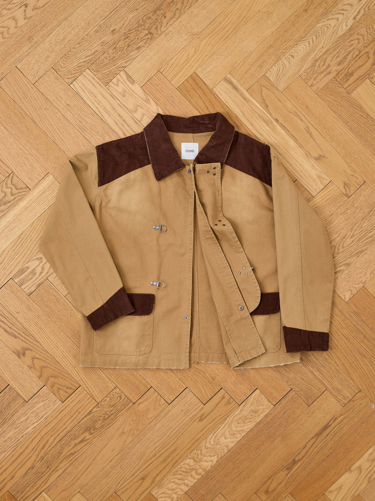 The Found Fireman Clasp Canvas Work Jacket, a unisex relaxed fit in tan washed canvas with dark brown collar and cuffs, lies flat on a light wood herringbone floor. Featuring sturdy metal clasps and a visible Found brand tag inside the collar, this mid-weight staple is perfect for everyday wear.