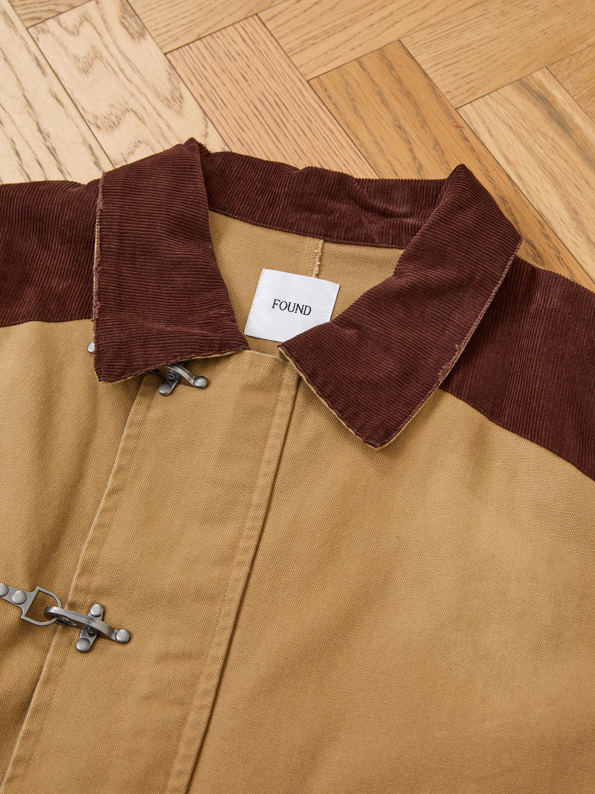 The Fireman Clasp Canvas Work Jacket by Found is crafted from tan washed canvas with dark brown corduroy detailing at the collar and shoulders, and features toggle fastenings and a distinctive white “FOUND” label. Designed as a mid-weight everyday staple, it offers a unisex relaxed fit for effortless style and comfort.