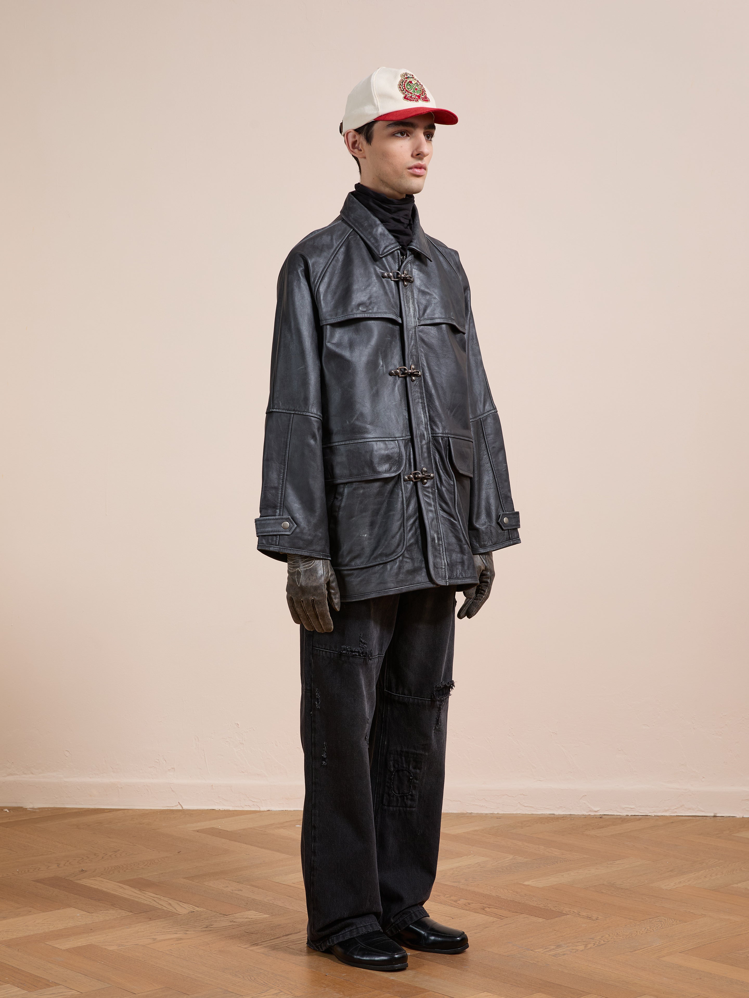 A person stands indoors on a wooden floor against a beige wall, wearing a white cap, black pants, gloves, shoes, and the FOUND Distressed Leather Fireman Clasp Jacket.