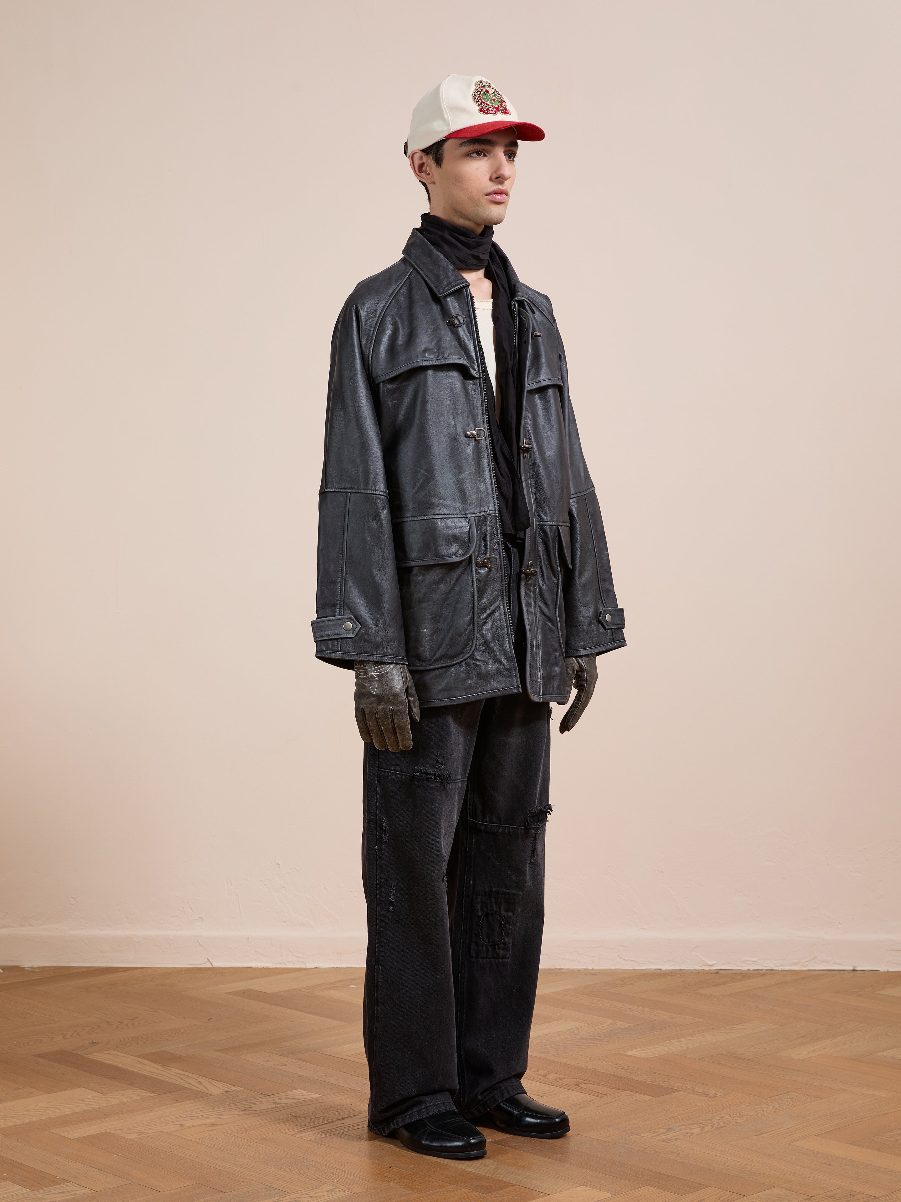 Indoors on a wooden floor, a person wears a white cap, black scarf, oversized FOUND Distressed Leather Fireman Clasp Jacket, dark wide-leg pants, gloves, and black shoes.