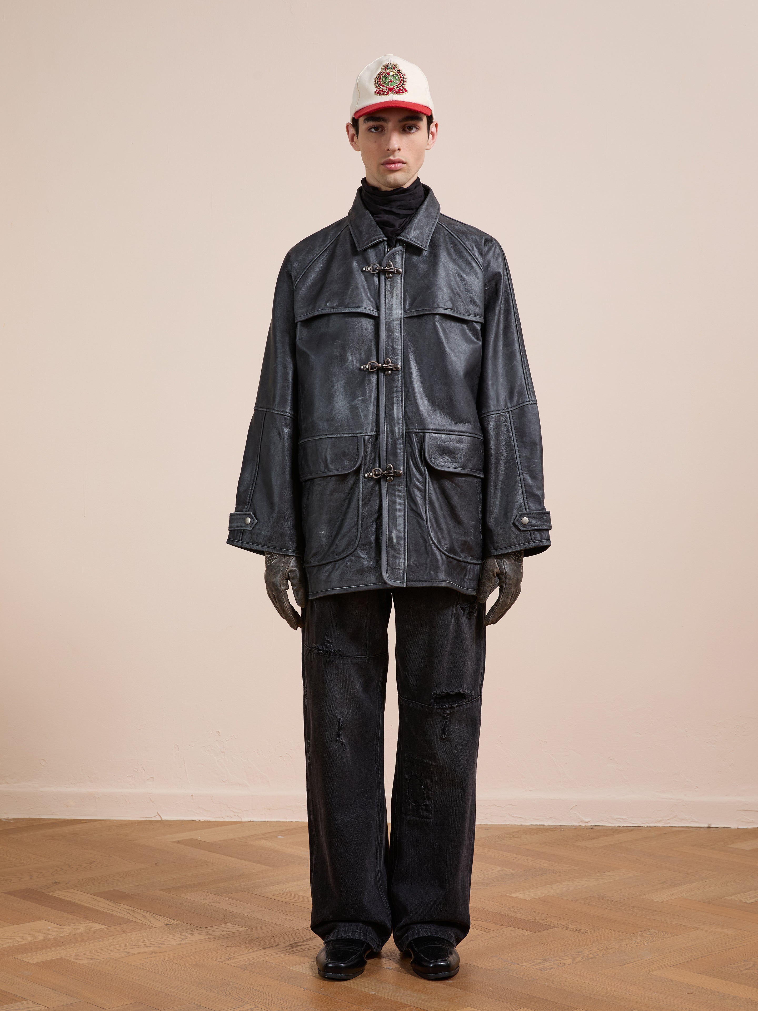 A person stands indoors facing forward against a plain beige wall, dressed in the FOUND Distressed Leather Fireman Clasp Jacket, black jeans, black gloves, and a white and red baseball cap.