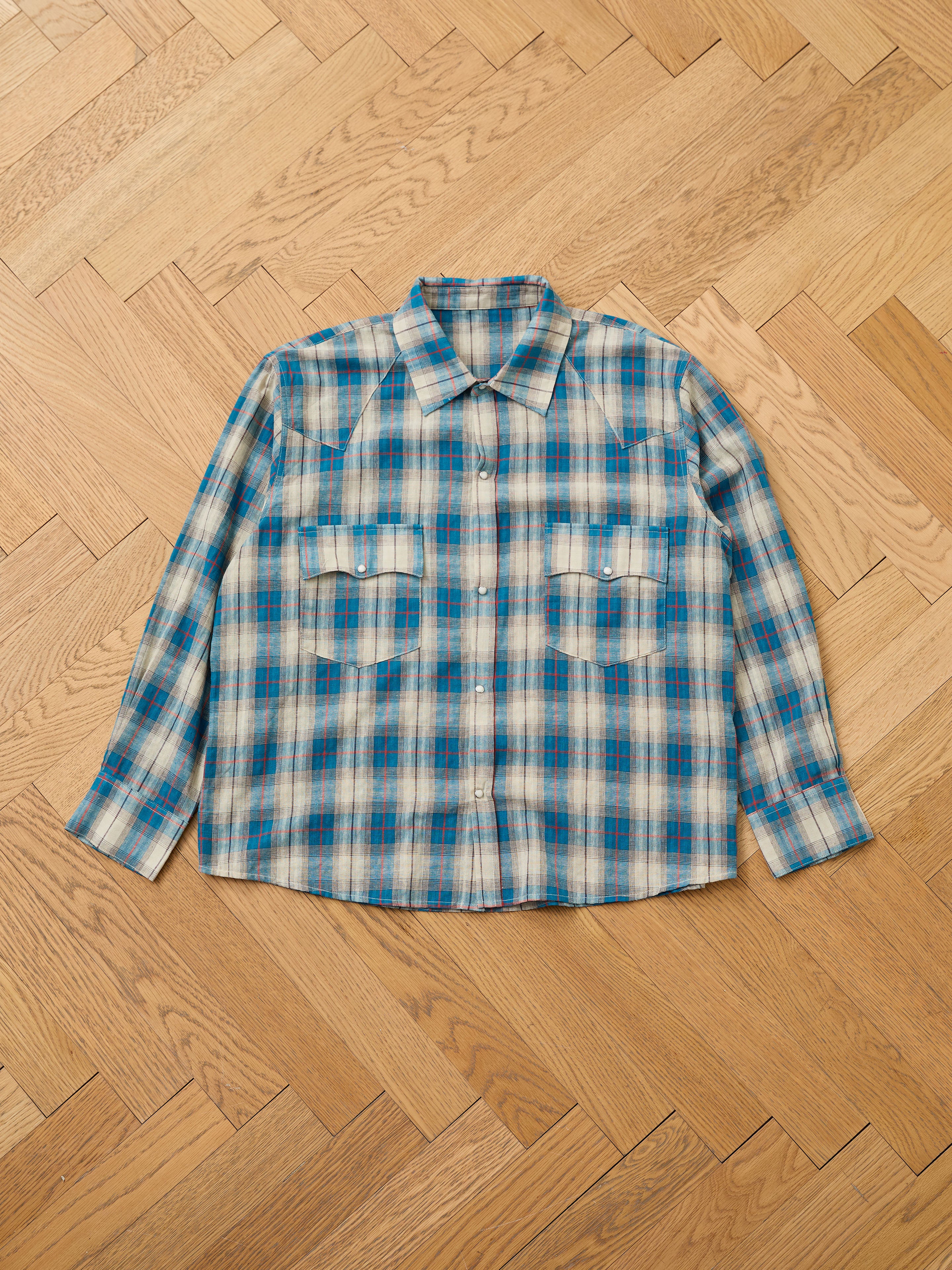 The FOUND Flannel Shirt is a unisex, relaxed fit button-up with long sleeves in blue, white, and beige plaid, shown laid flat on a light wooden herringbone floor—a classic wardrobe addition.