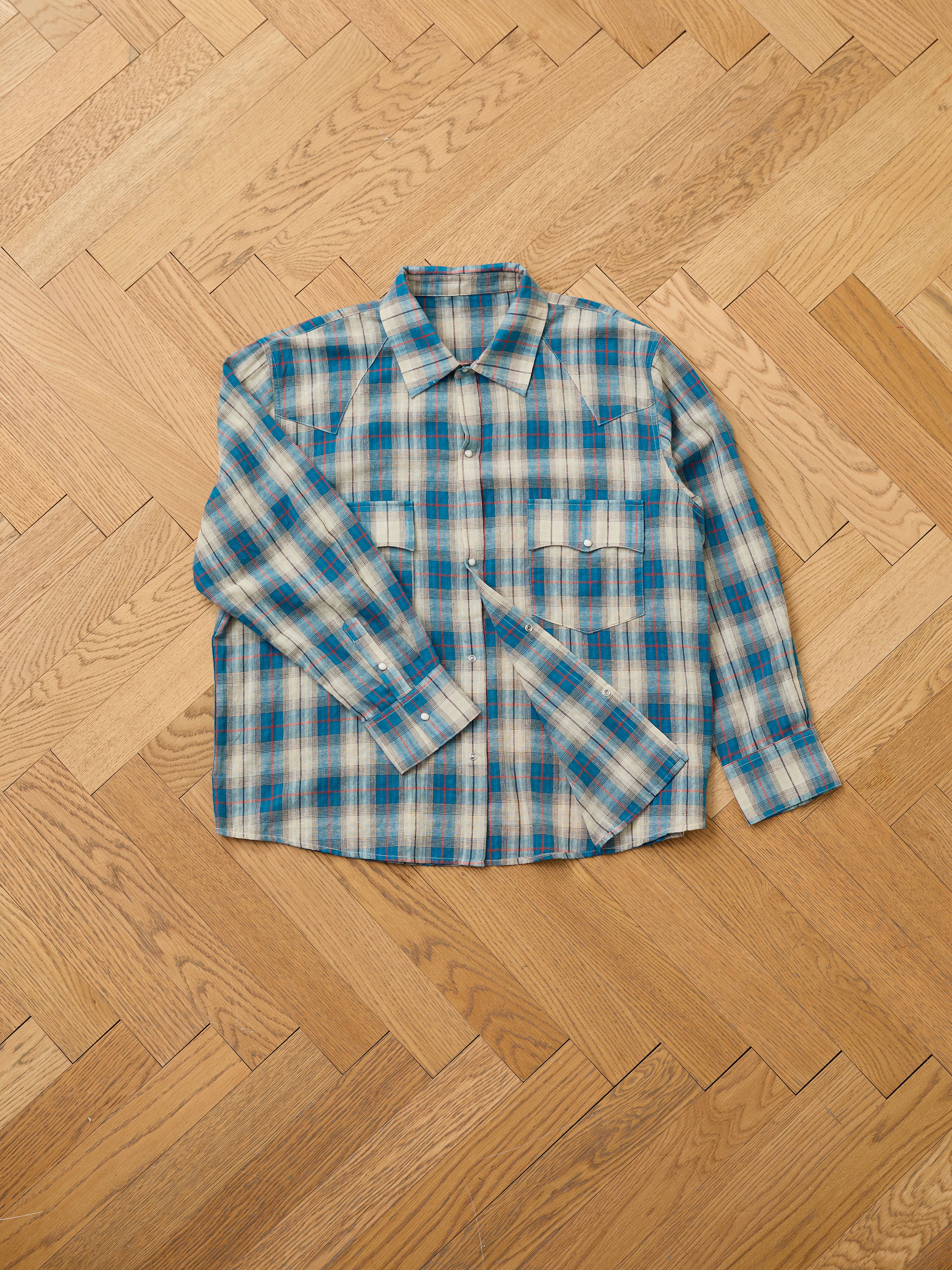 The FOUND Flannel Shirt, featuring a blue and beige Western plaid design and a unisex relaxed fit, is displayed flat on a light wooden herringbone floor.