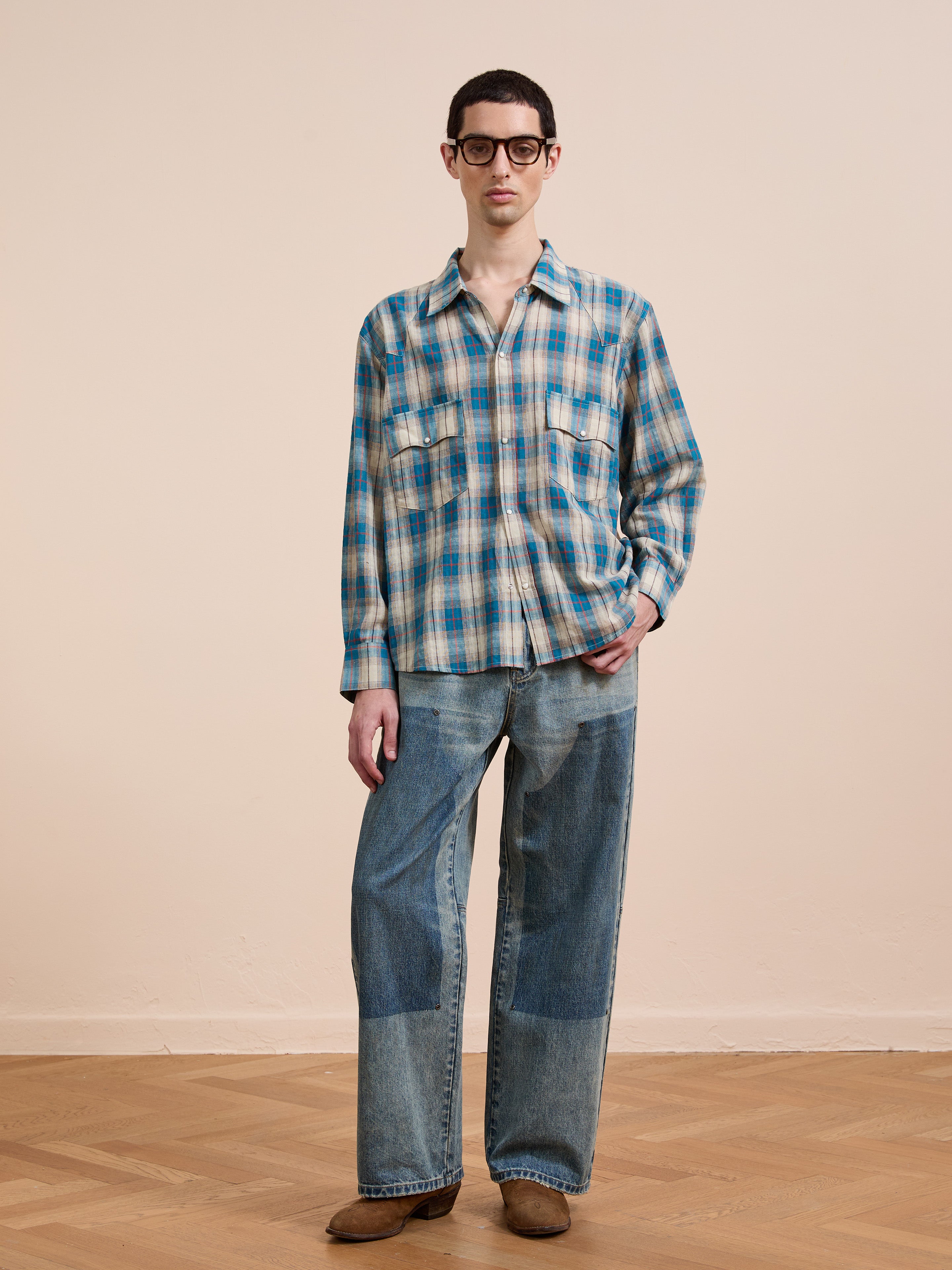 A person wearing glasses, a FOUND Flannel Shirt in a unisex relaxed fit, wide-leg patchwork jeans, and brown shoes stands on a wooden floor against a plain beige wall.