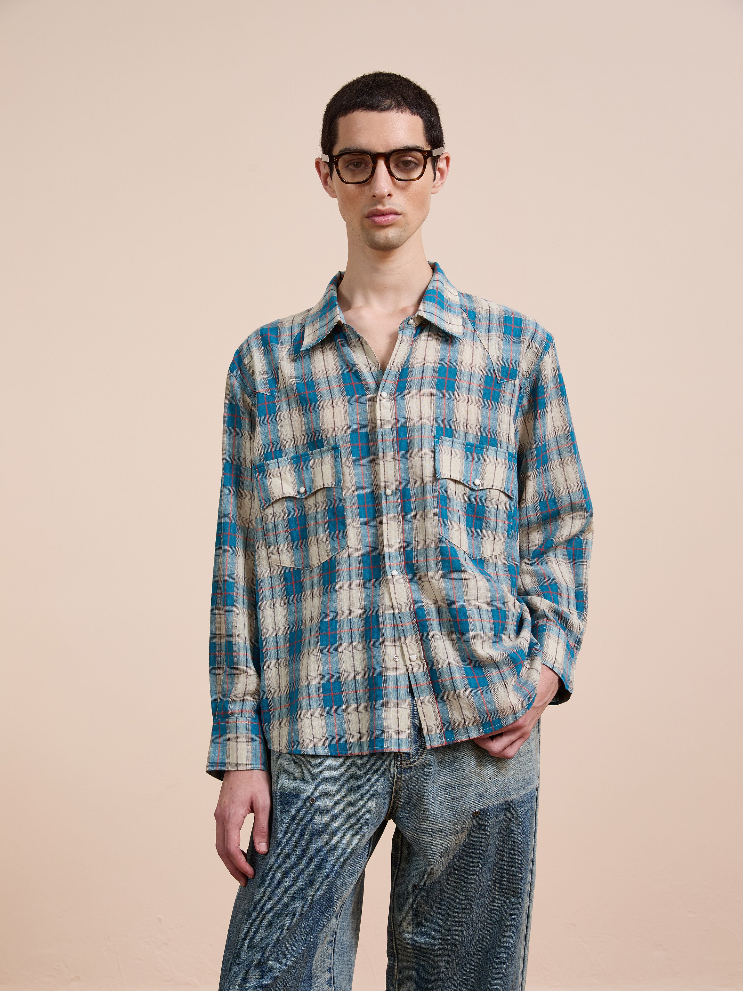 A person with short dark hair and glasses wears the FOUND Flannel Shirt in a unisex relaxed fit, featuring blue and beige Western plaid, paired with light blue jeans against a plain beige background.