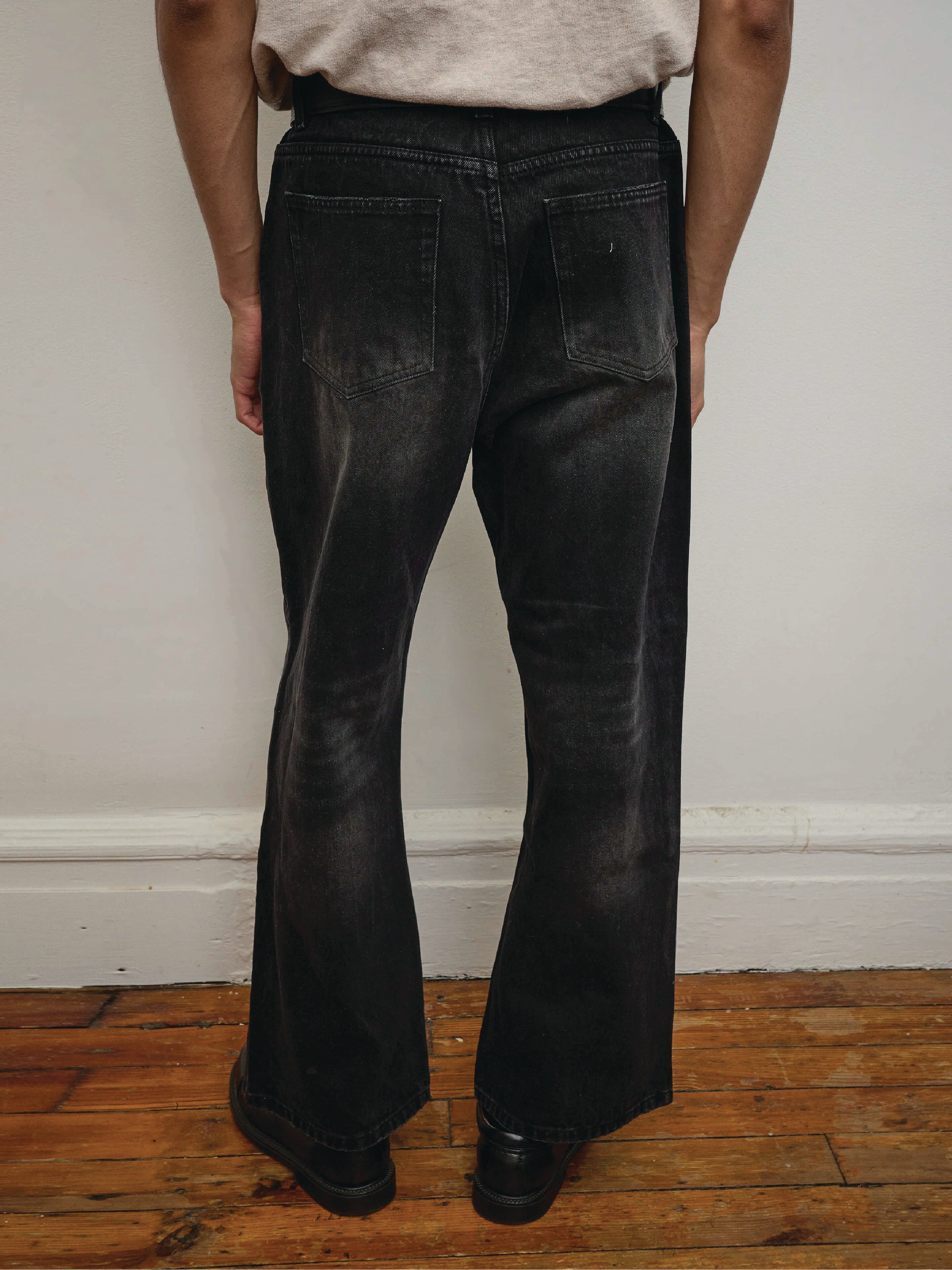 A person wearing Found Flare Jeans in a vintage black wash and black shoes stands on a wooden floor against a white wall, shown from the back.