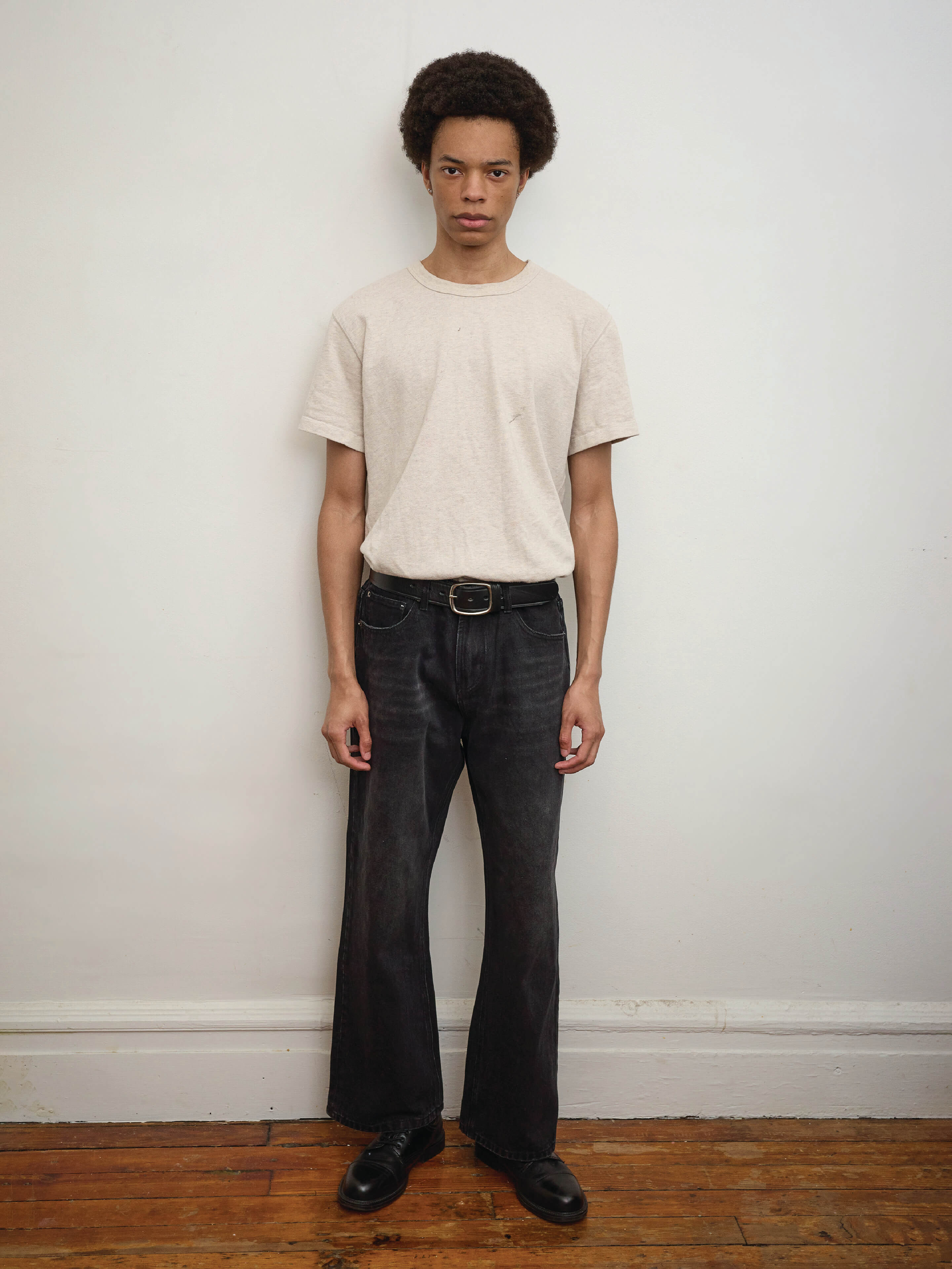 A person with an afro stands against a white wall, wearing a beige t-shirt, Found Flare Jeans in vintage black wash, and black shoes on a wooden floor.