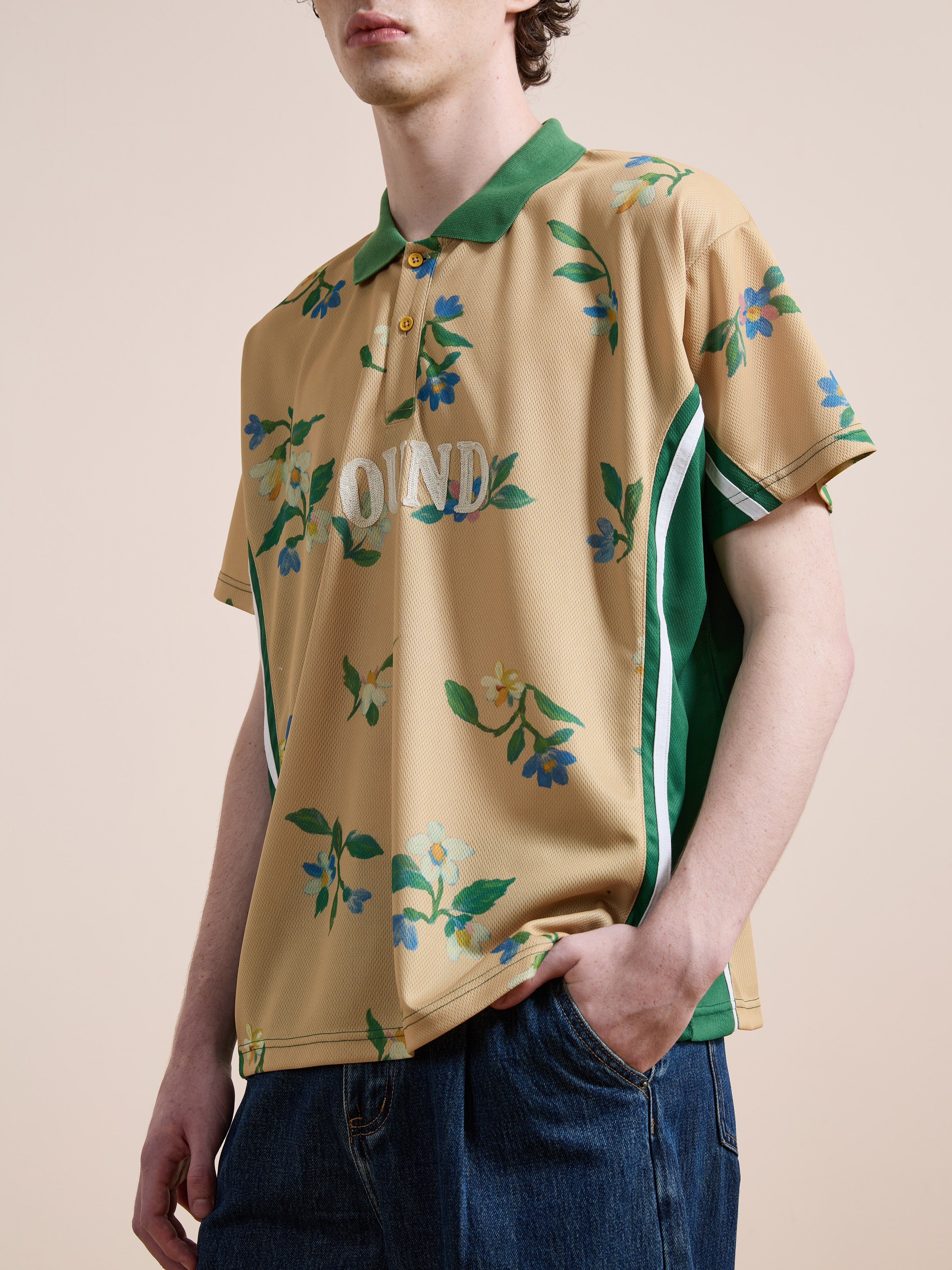 A person wearing the FOUND Floral Polo Jersey—tan with botanical embroidery, green collar and side panels—paired with blue jeans stands against a plain background.