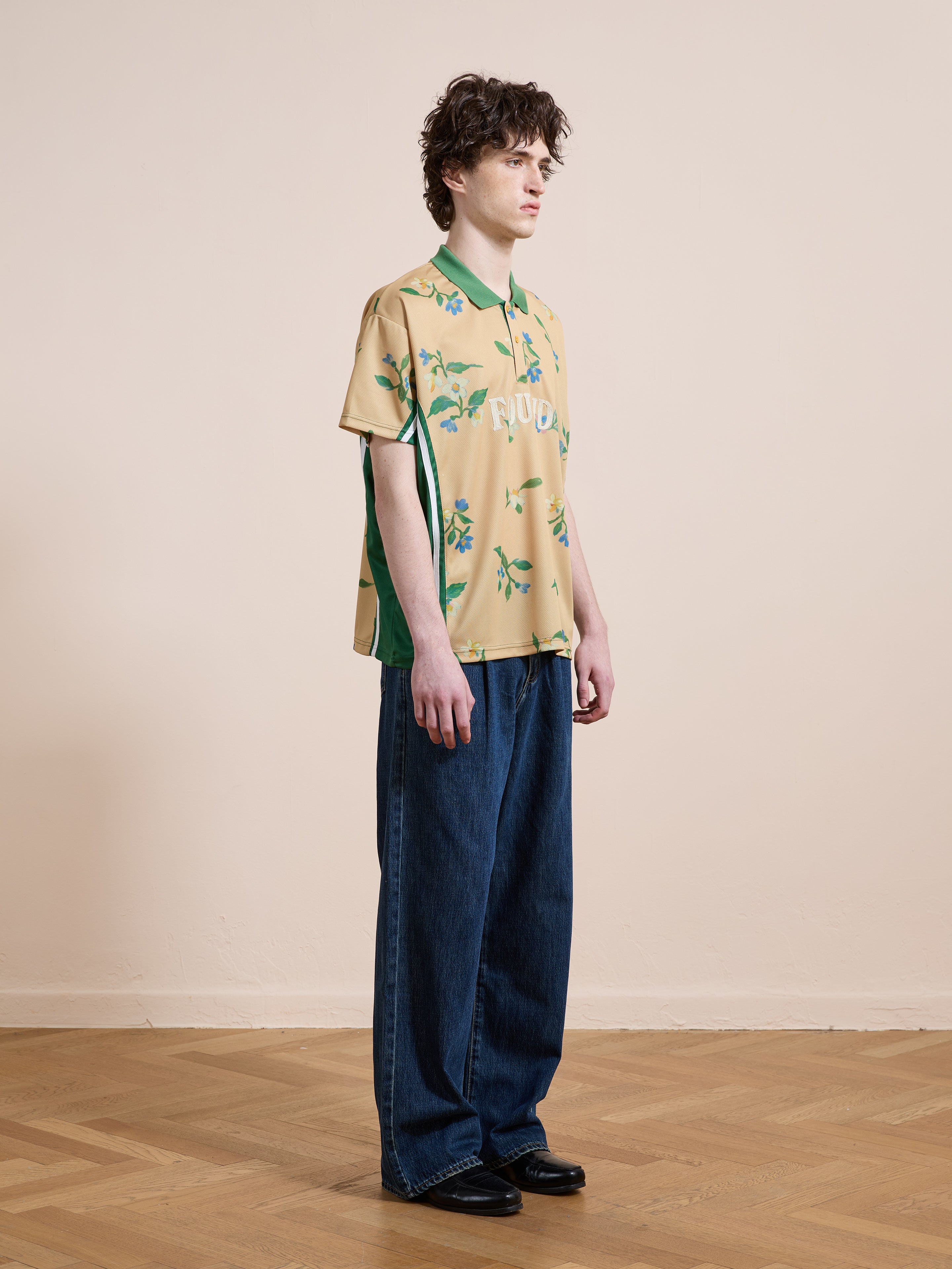 A person stands indoors wearing the FOUND Floral Polo Jersey, a unisex relaxed fit top, paired with loose blue jeans and black shoes against a plain light-colored wall and wooden floor.
