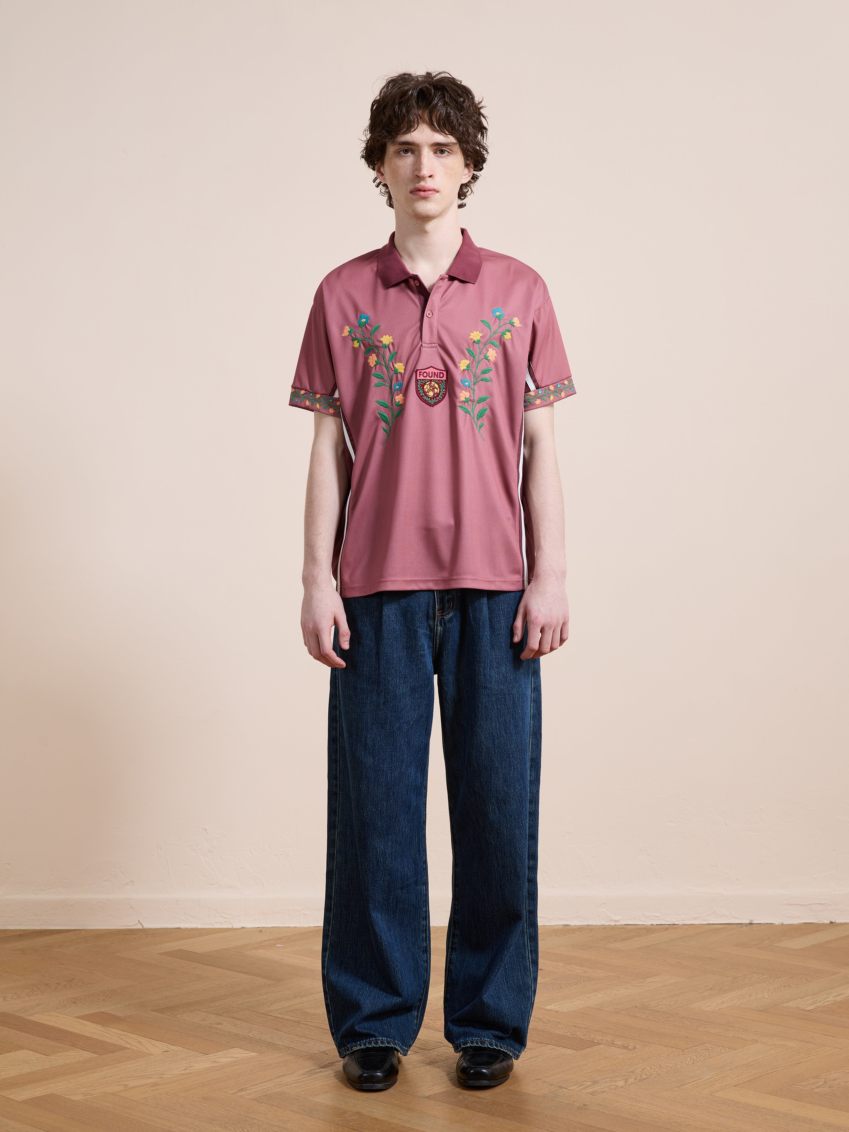 A person stands indoors, facing forward, wearing the FOUND Floral Polo Jersey and wide-leg blue jeans on a wooden floor against a plain light wall.