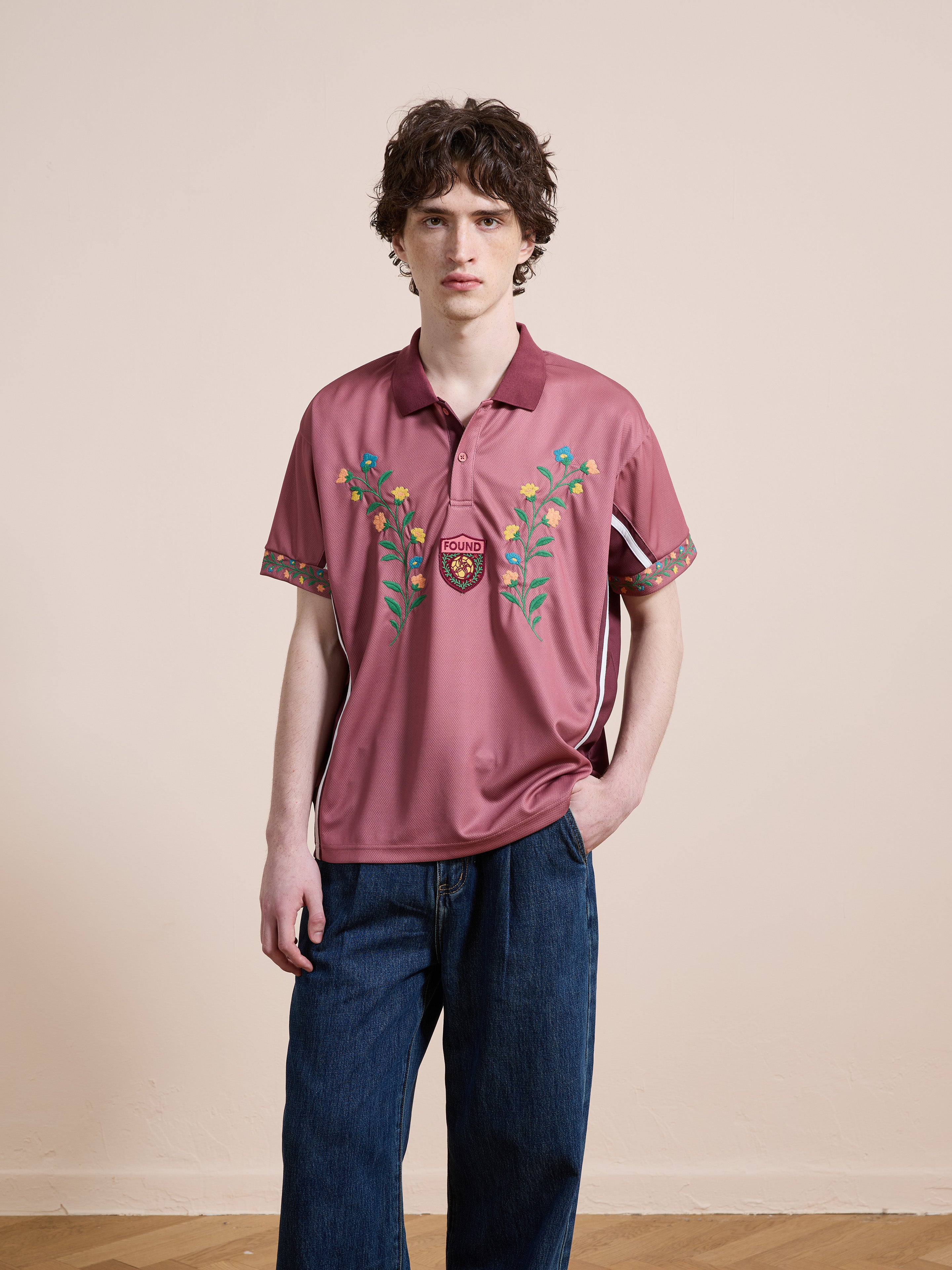 A person is indoors wearing the FOUND Floral Polo Jersey and wide-leg blue jeans, standing against a plain beige wall.