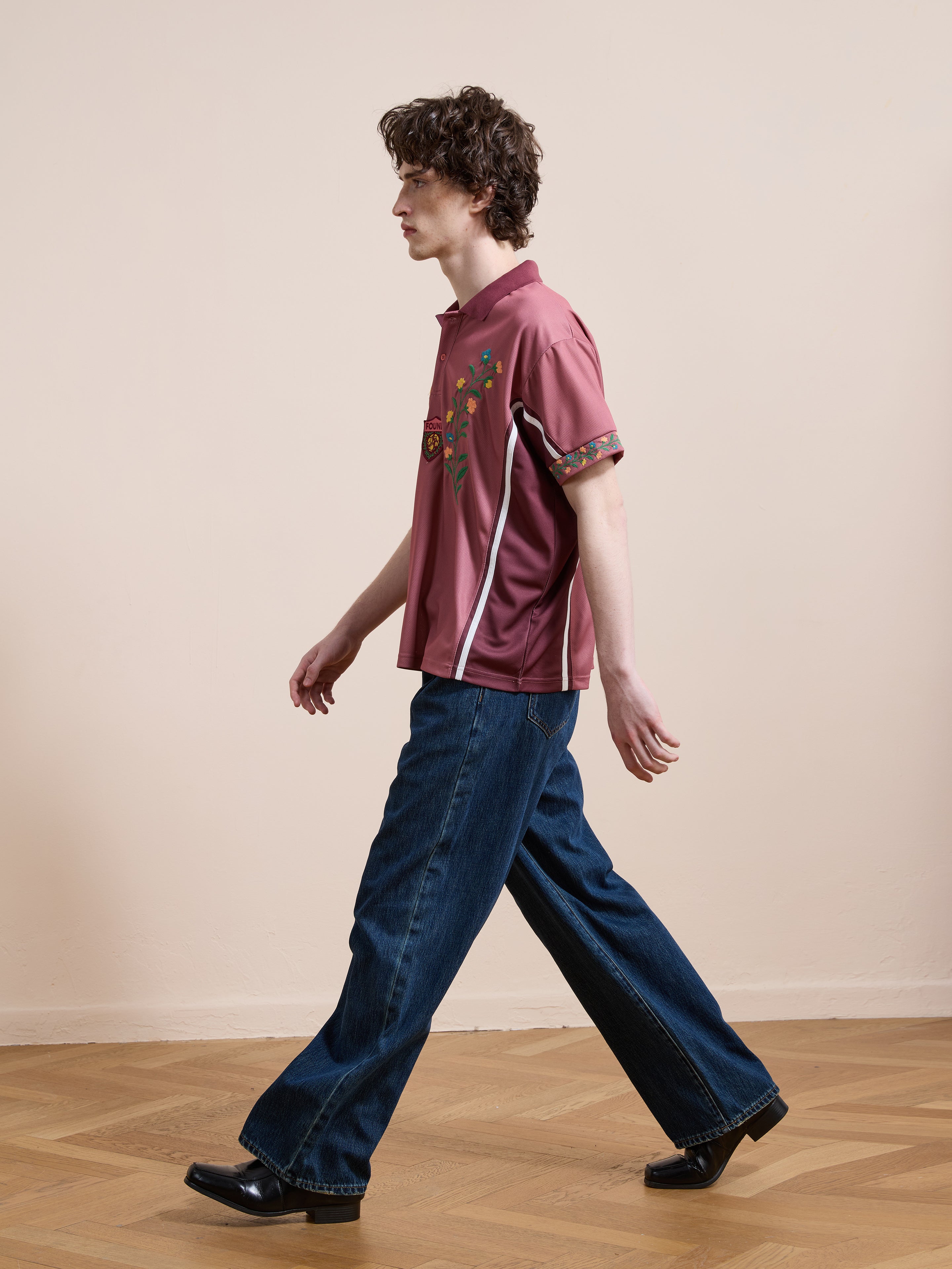 A male model walks in profile wearing the FOUND Floral Polo Jersey, wide-leg dark jeans, and black boots on a herringbone wood floor.