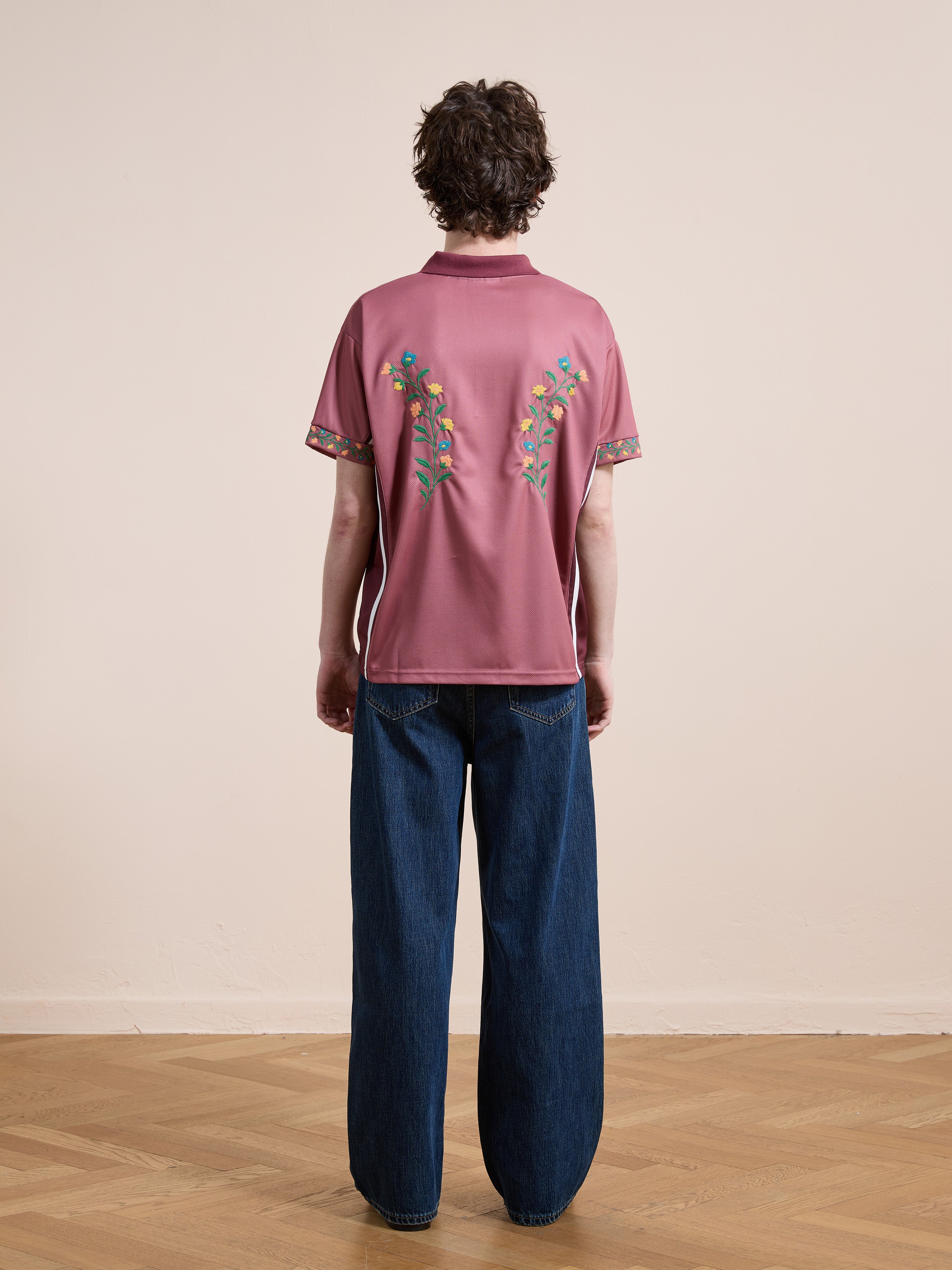Person standing indoors facing away, wearing FOUND's Floral Polo Jersey—a mauve cotton unisex top with floral embroidery on the back—and loose-fitting blue jeans, on a wooden floor against a plain light wall.