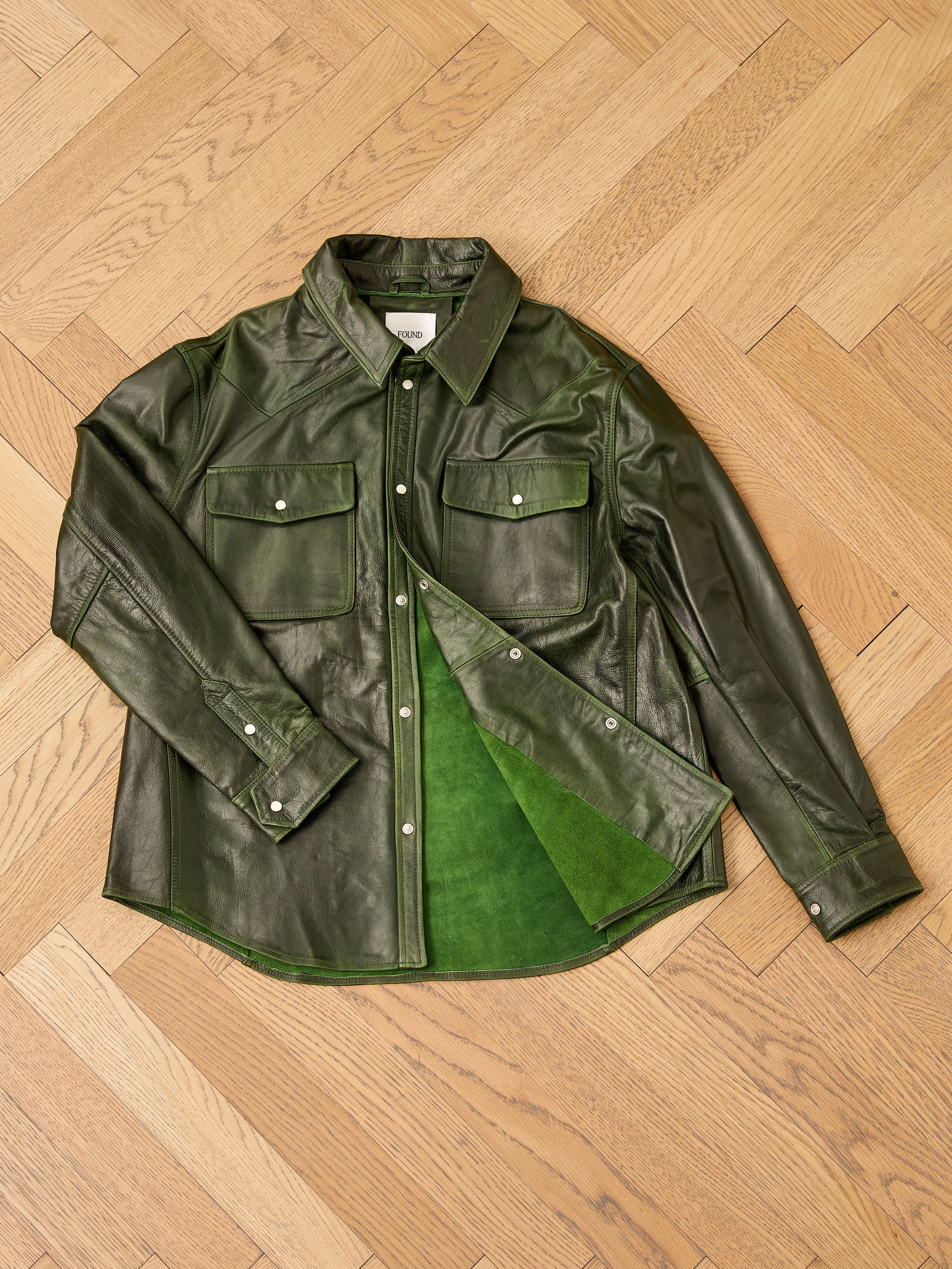 The FOUND Forest Leather Western Shirt, featuring snap buttons and two chest pockets, is laid flat on a wooden herringbone floor with its front partially unbuttoned to reveal the rich green lining.