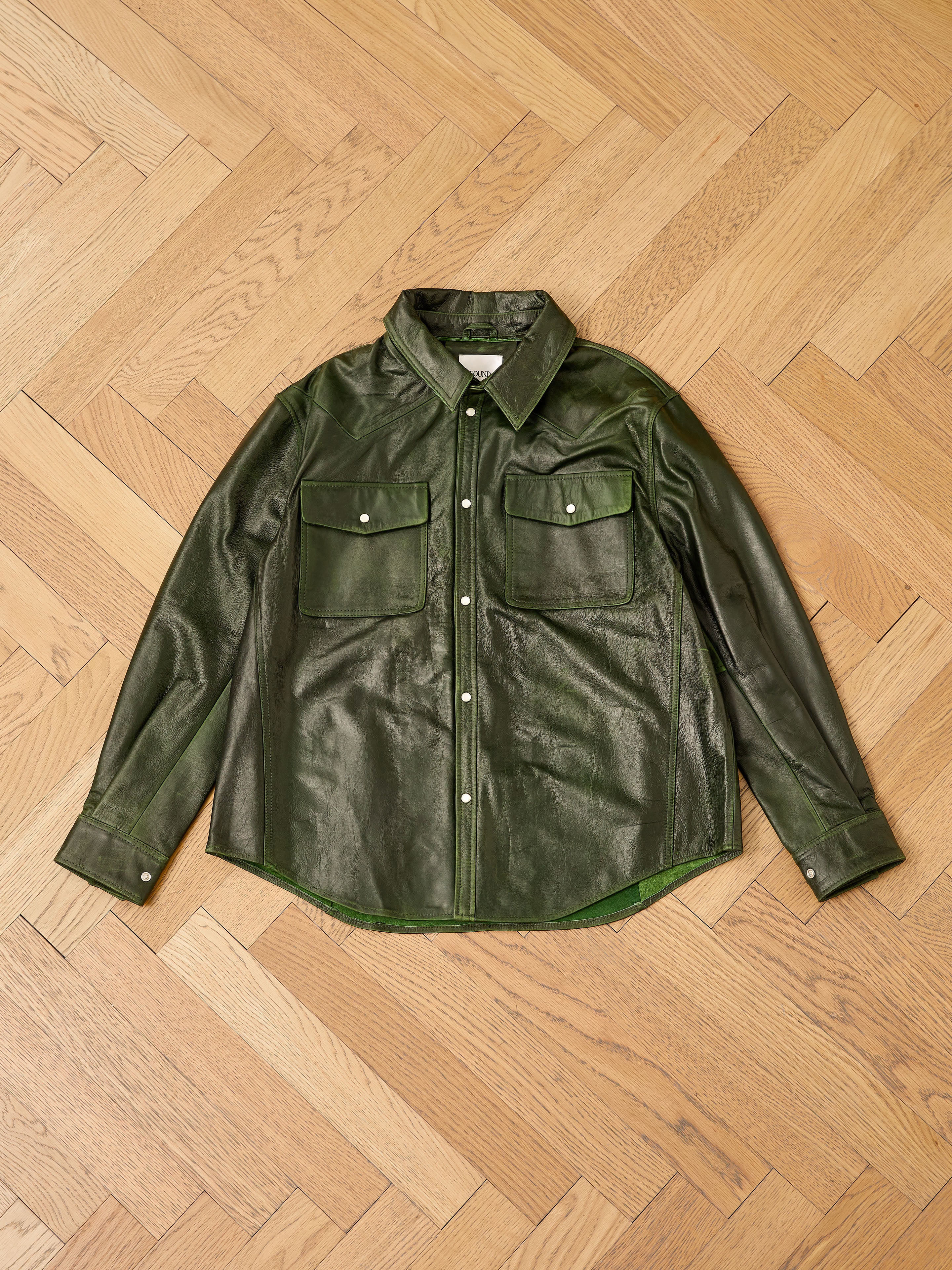 The FOUND Forest Leather Western Shirt, featuring a unisex western style in green leather, rests on a wooden floor, adding rugged charm to the scene.