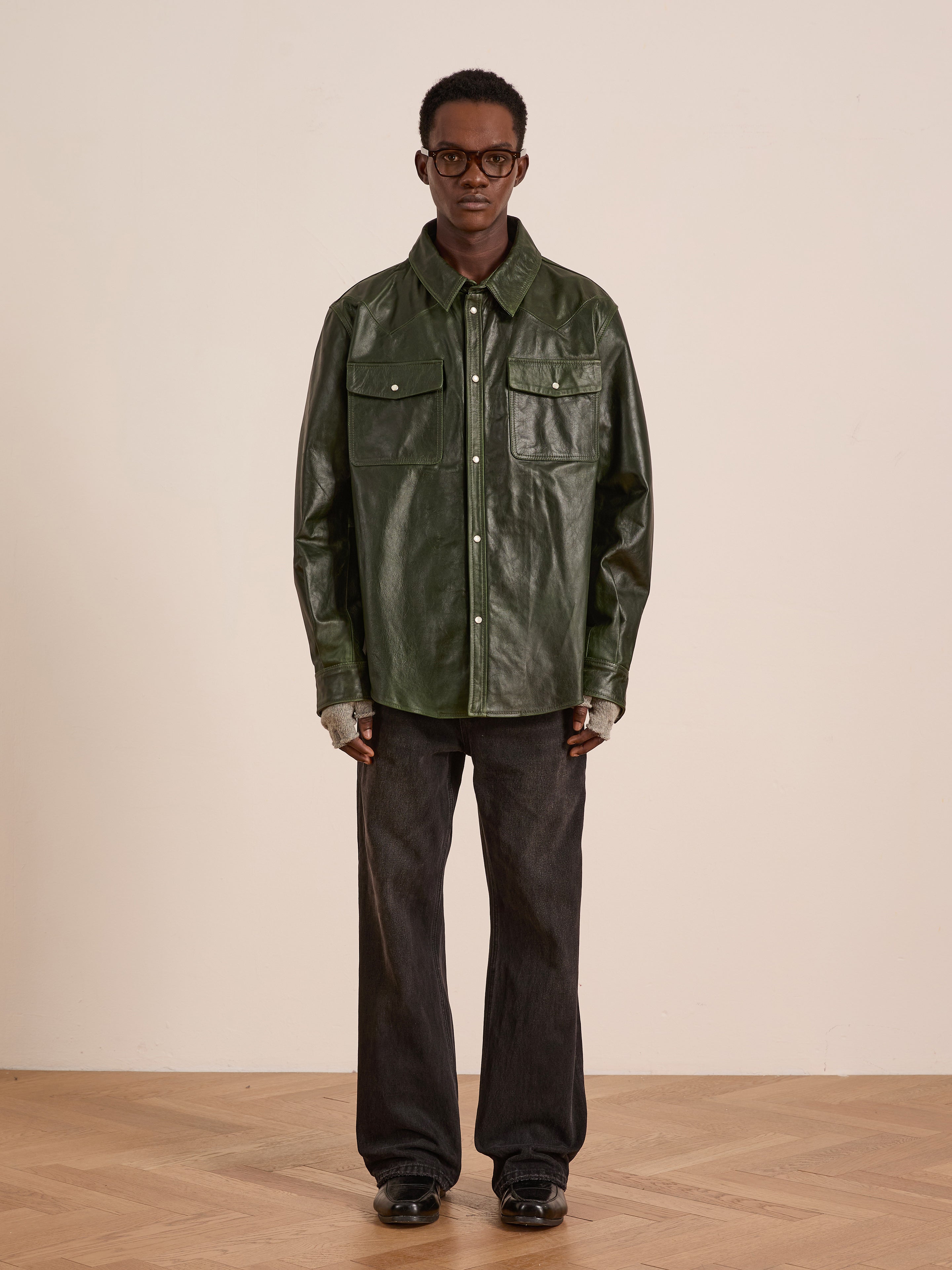 Indoors against a plain light wall, a person wears the FOUND Forest Leather Western Shirt, paired with dark wide-leg pants, black shoes, glasses, and fingerless gloves.