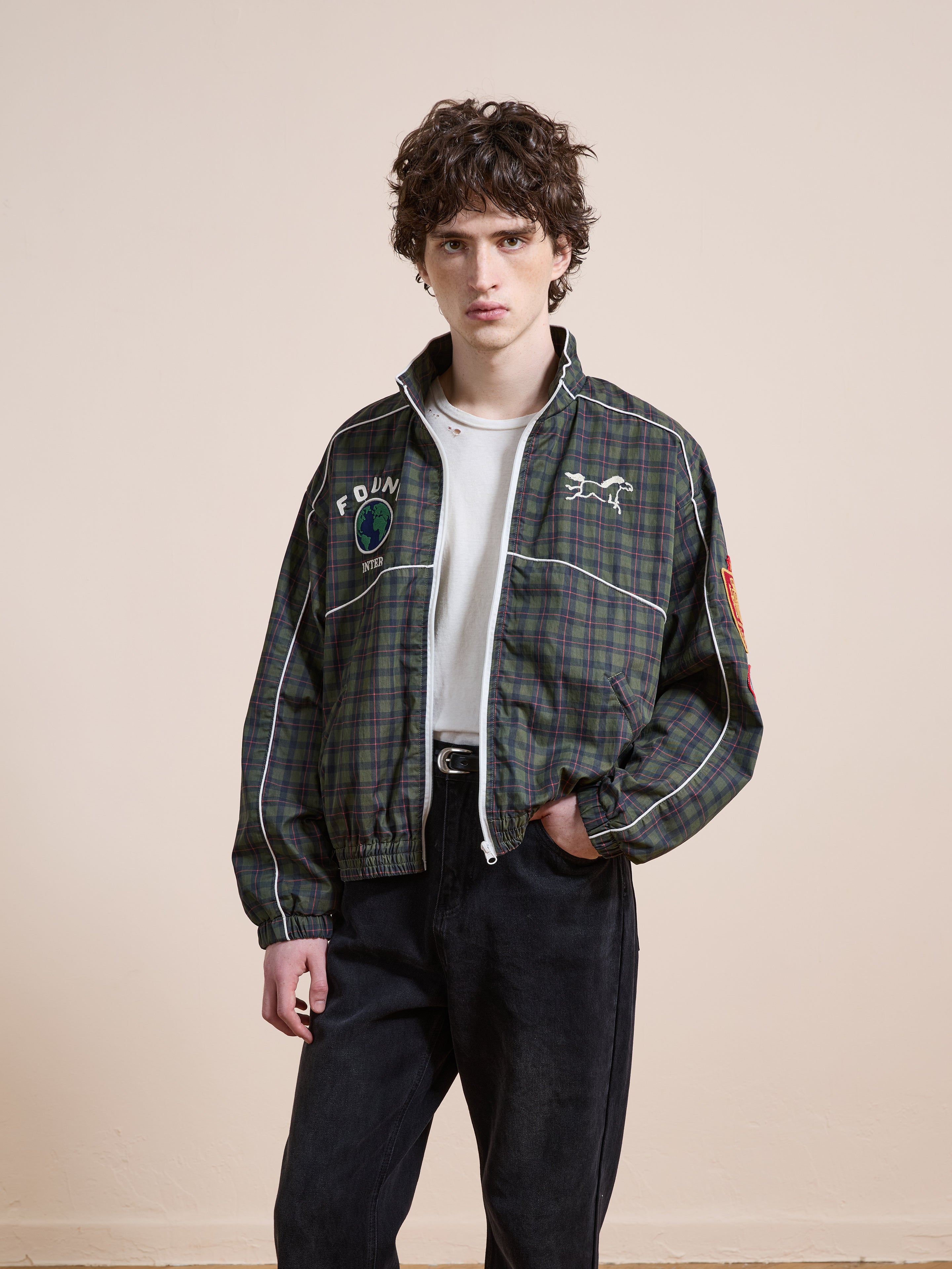 A person stands against a plain background wearing the FOUND Forest Plaid Track Jacket, paired with a white t-shirt and black pants.