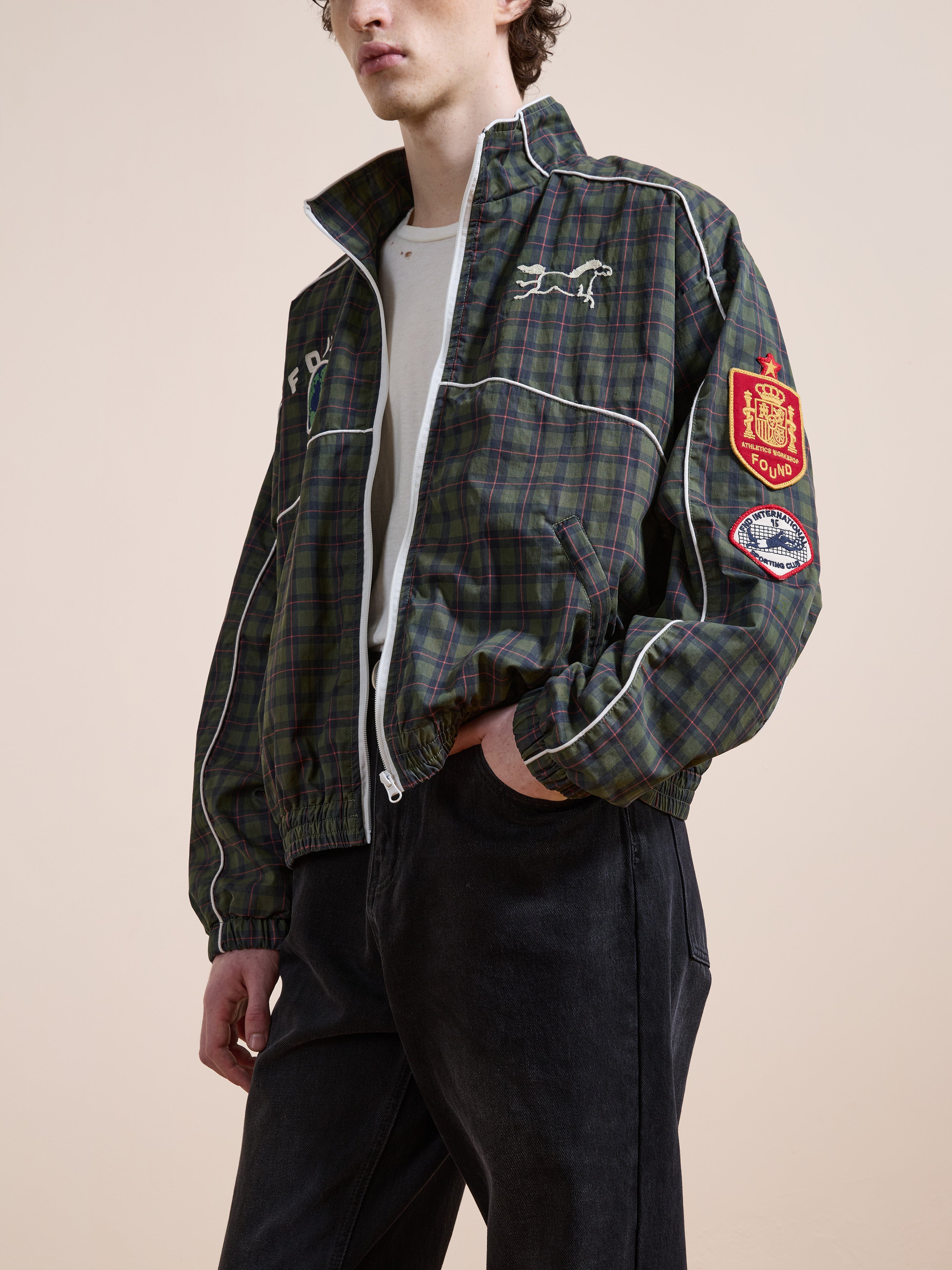 A person wears the FOUND Forest Plaid Track Jacket, featuring white piping, embroidered logos, and sleeve patches, styled with a white shirt and black pants.