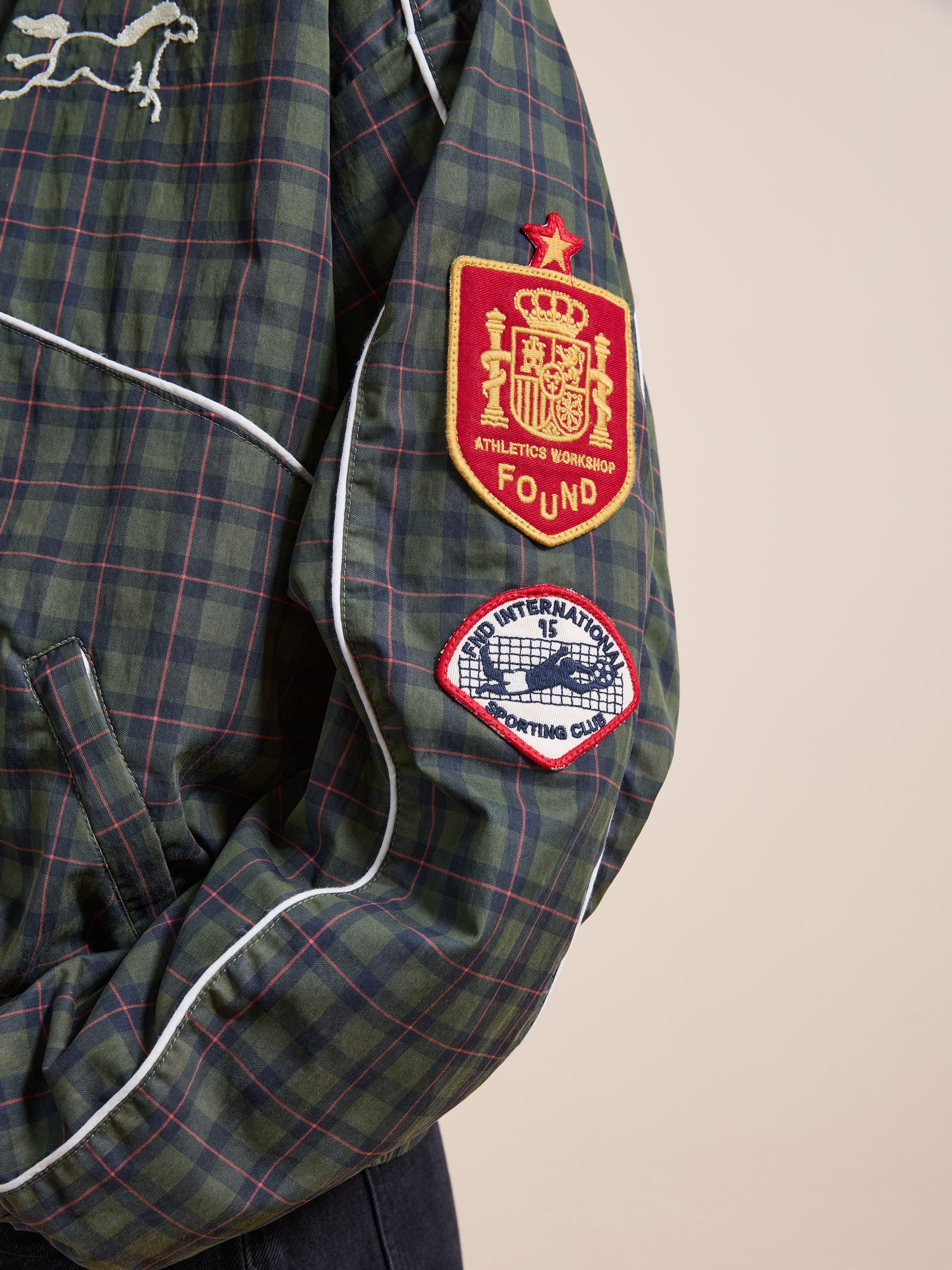 A close-up of the FOUND Forest Plaid Track Jacket sleeve features two embroidered patches: a red and gold crest and a white and blue circular patch.