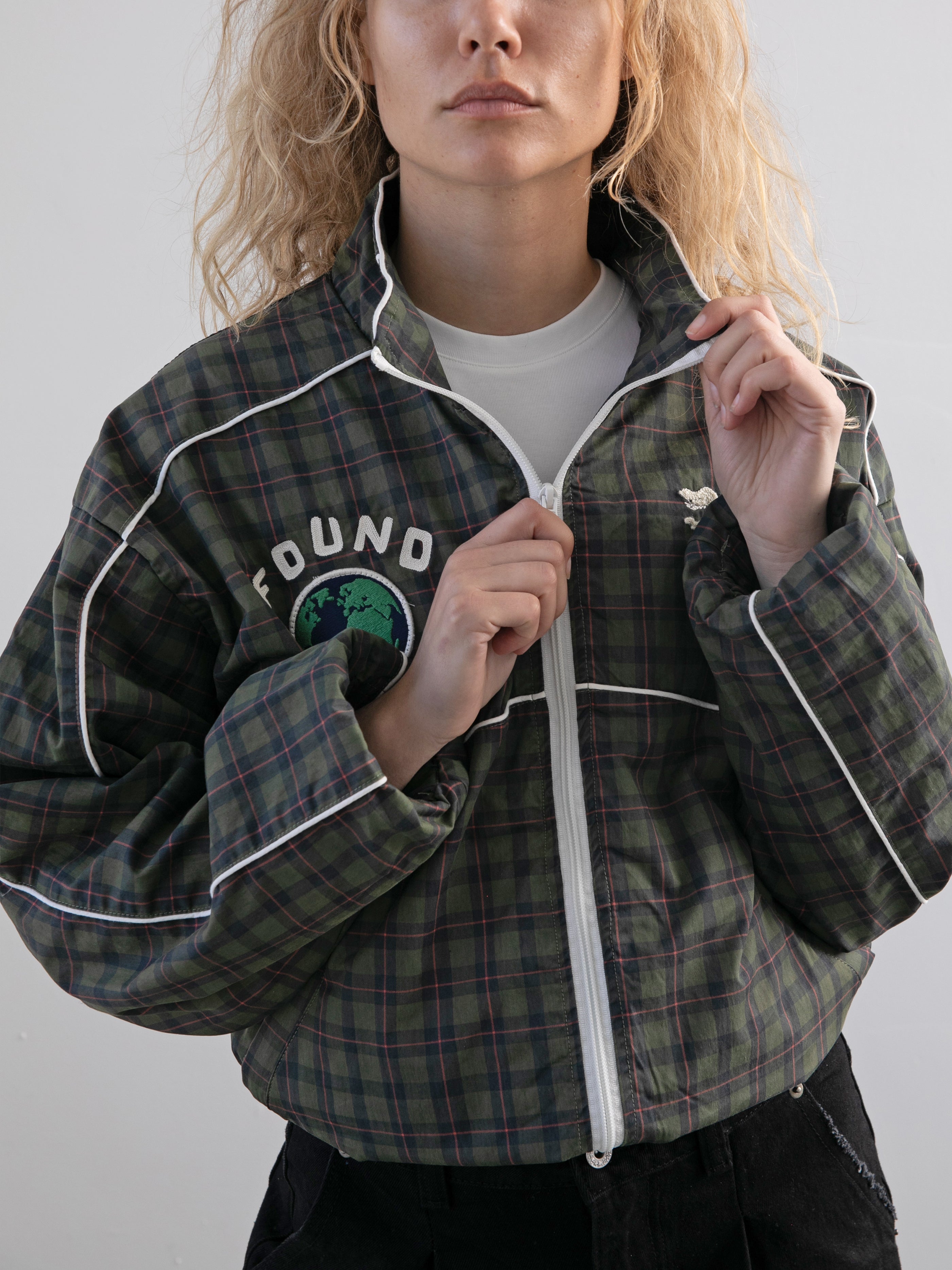 A person with wavy blonde hair zips up the FOUND Forest Plaid Track Jacket, a green plaid style with white piping, "FOUND" lettering, and an earth graphic on the chest.