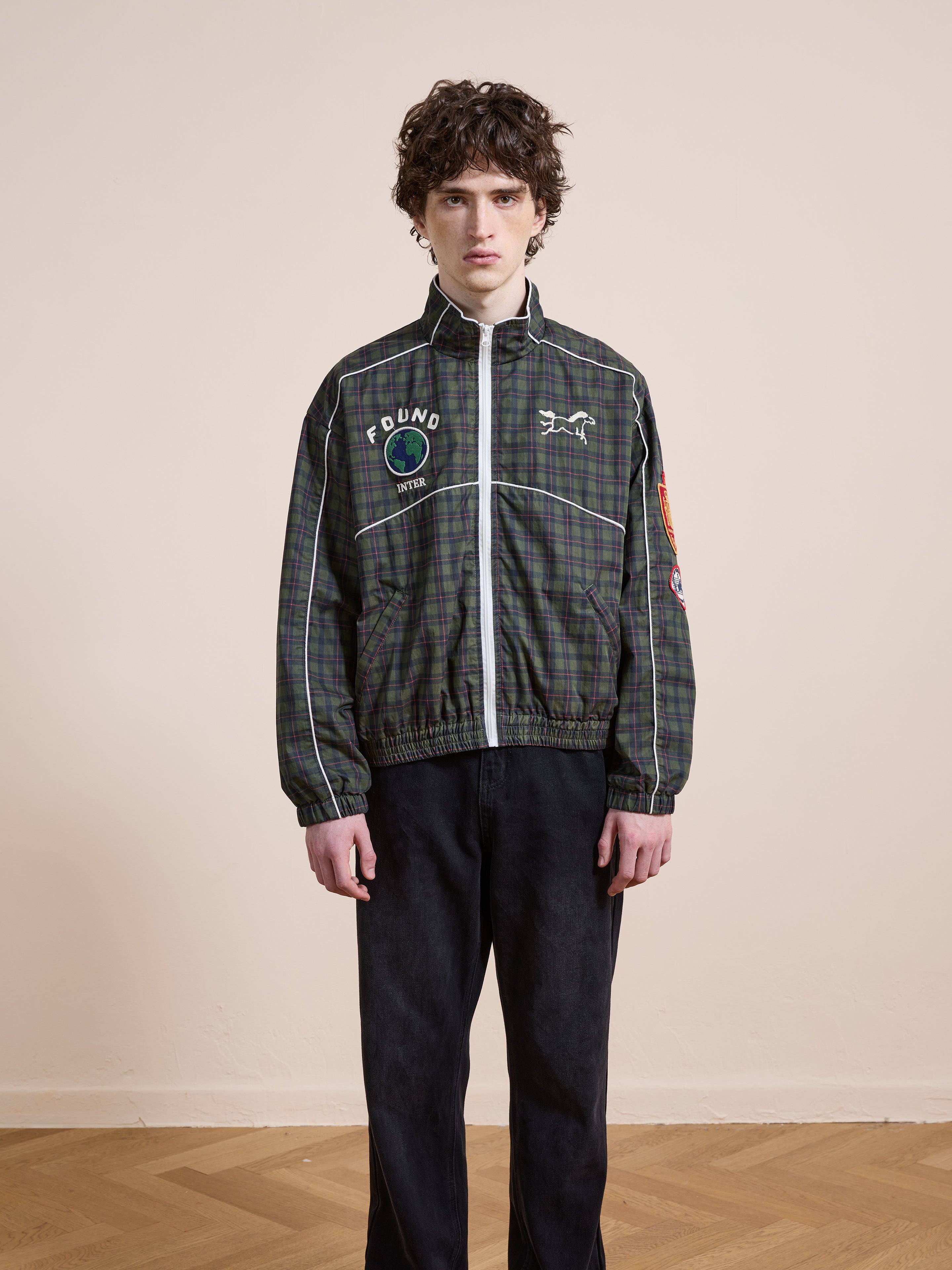 Indoors, a person wears the FOUND Forest Plaid Track Jacket featuring vintage plaid, globe and text patches, styled with black pants.