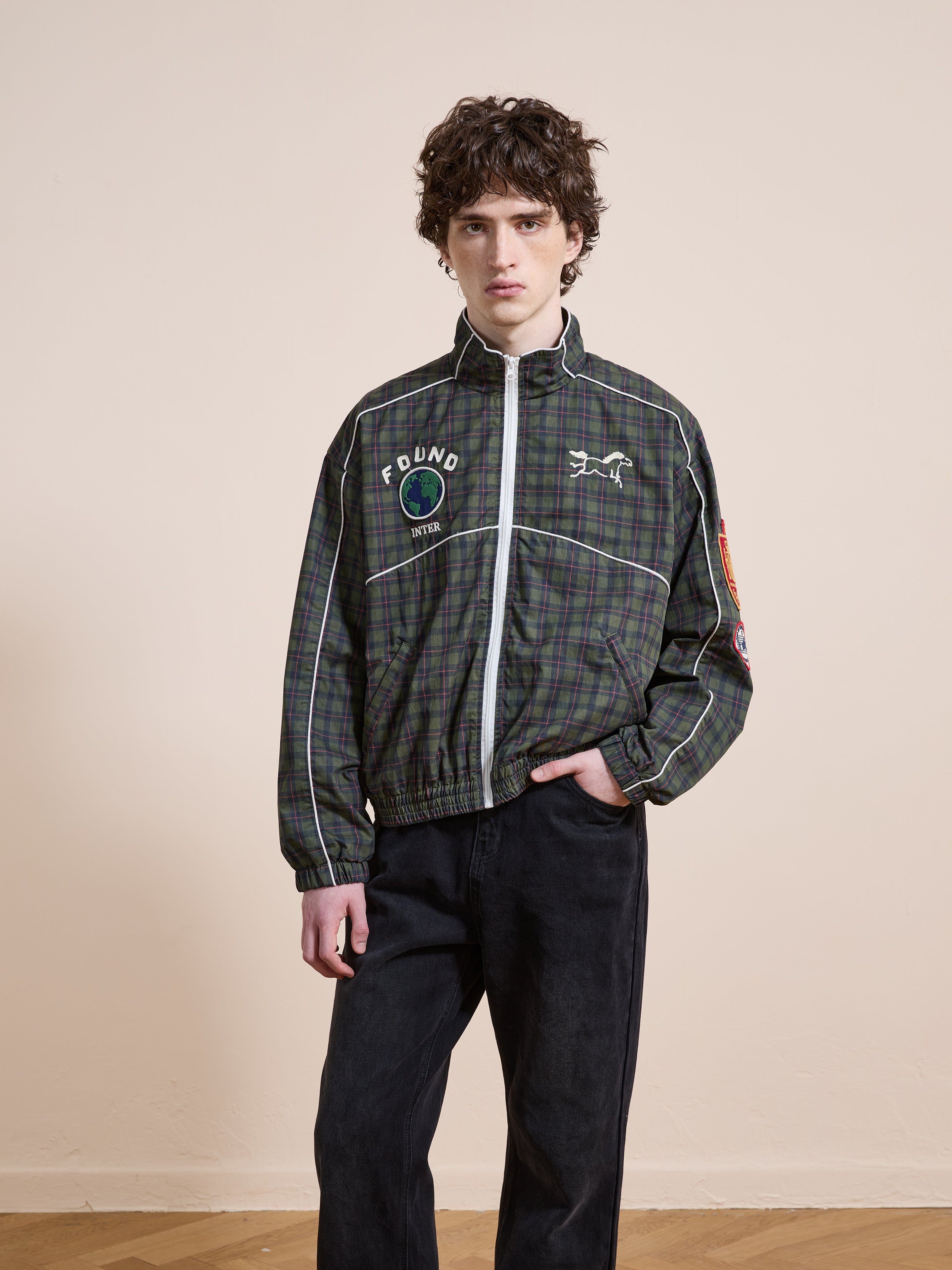 A person with curly hair wears the FOUND Forest Plaid Track Jacket with black pants, standing indoors against a plain light-colored wall.