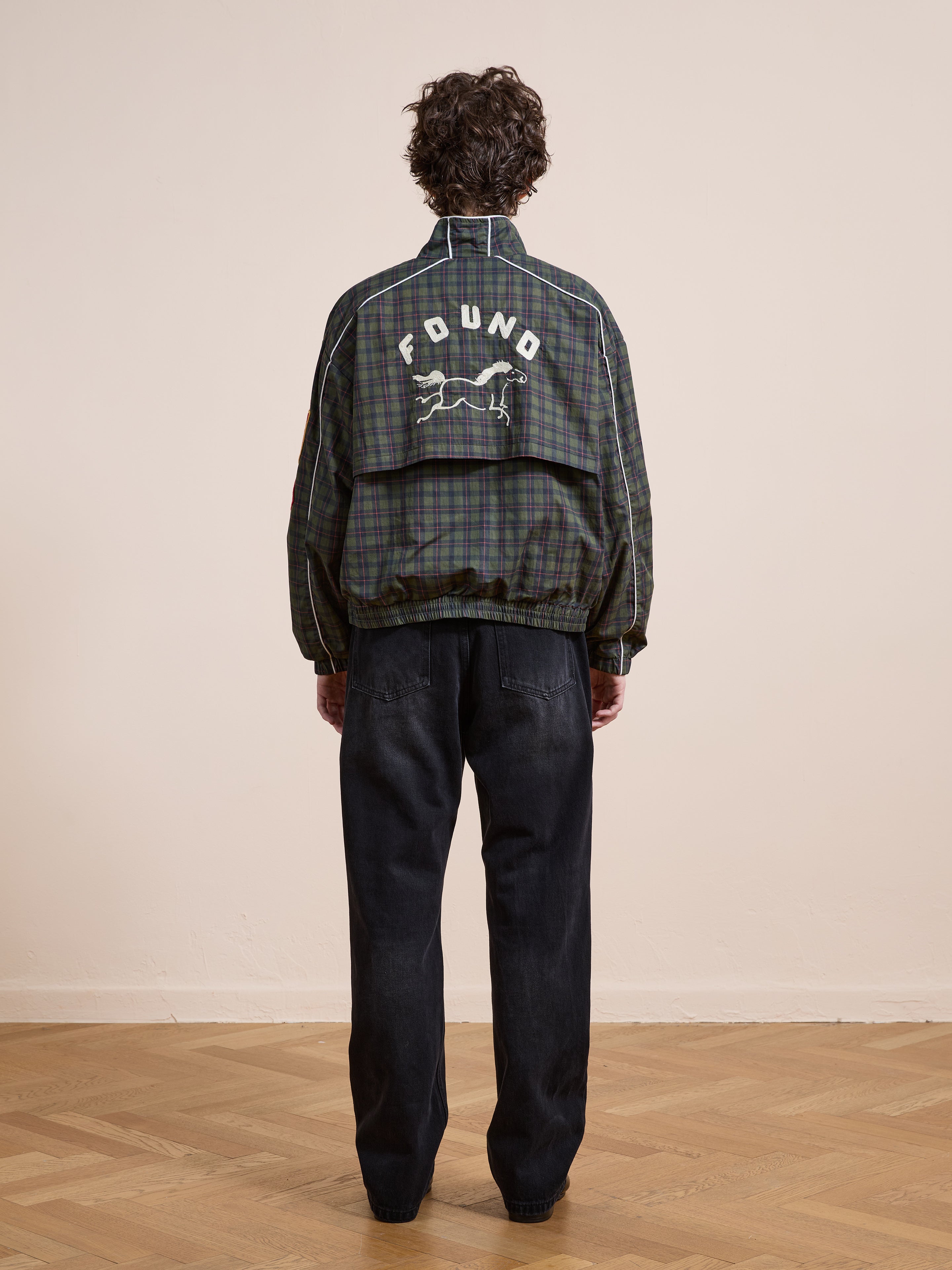 A person stands indoors on a wooden floor, facing away, wearing the FOUND Forest Plaid Track Jacket with "FOUND" and a white horse graphic on the back, paired with black jeans.
