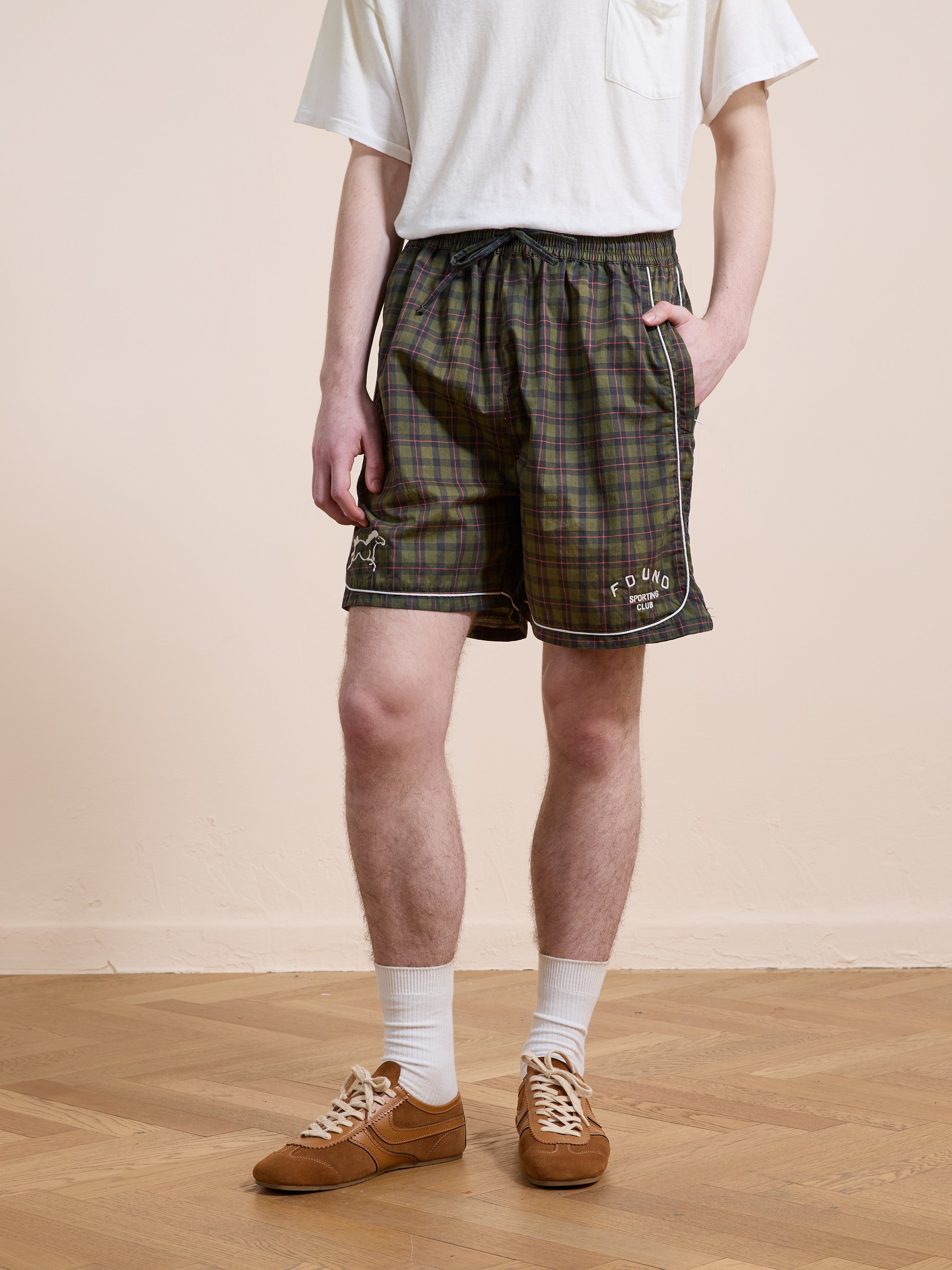 A person in a white T-shirt, Found Forest Plaid Track Shorts (unisex, relaxed fit), white socks, and brown sneakers stands on a wooden floor against a plain light-colored wall.