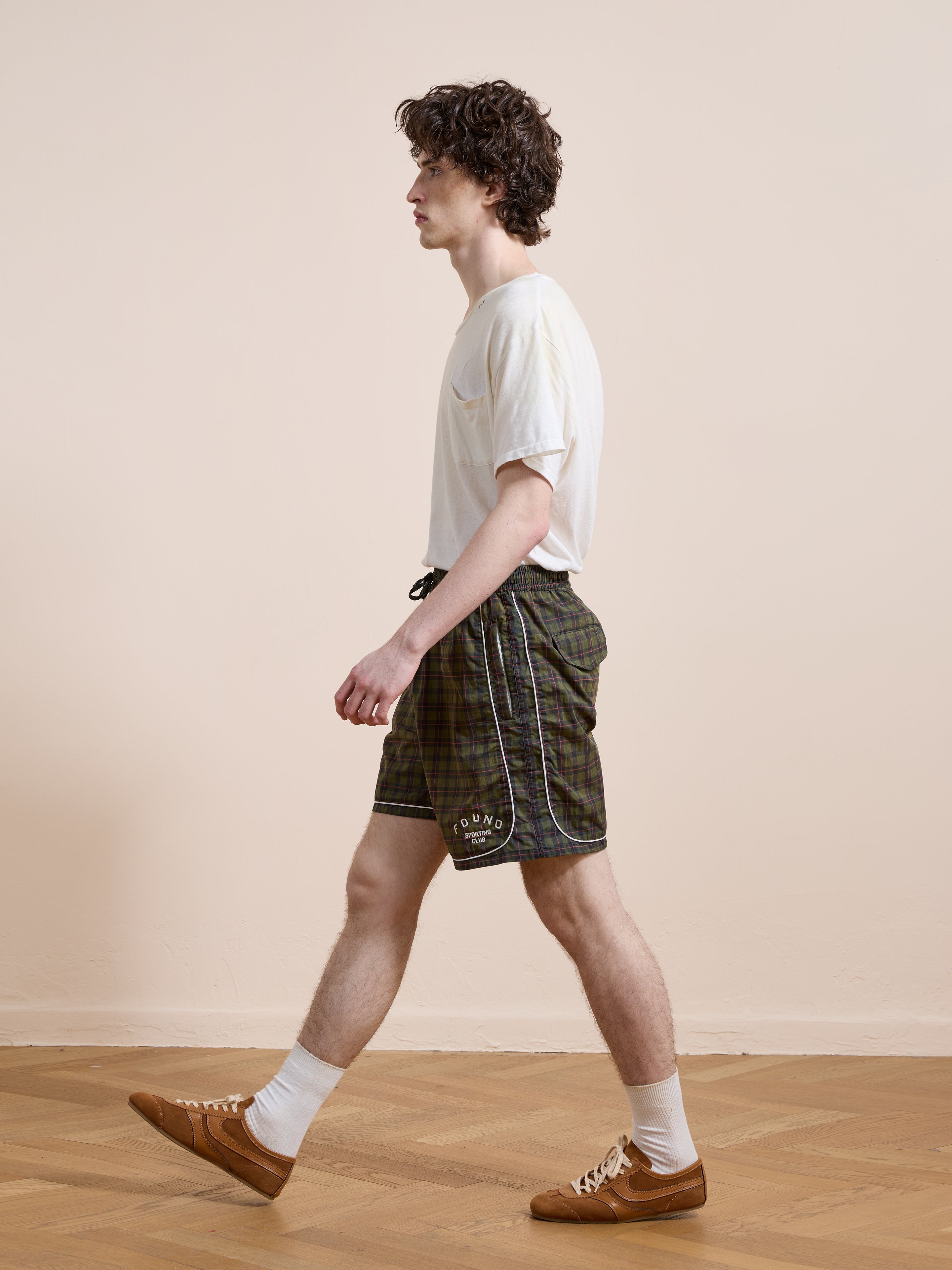 A man in a white t-shirt, Found Forest Plaid Track Shorts, white socks, and brown sneakers walks indoors on a wooden floor against a plain beige wall.