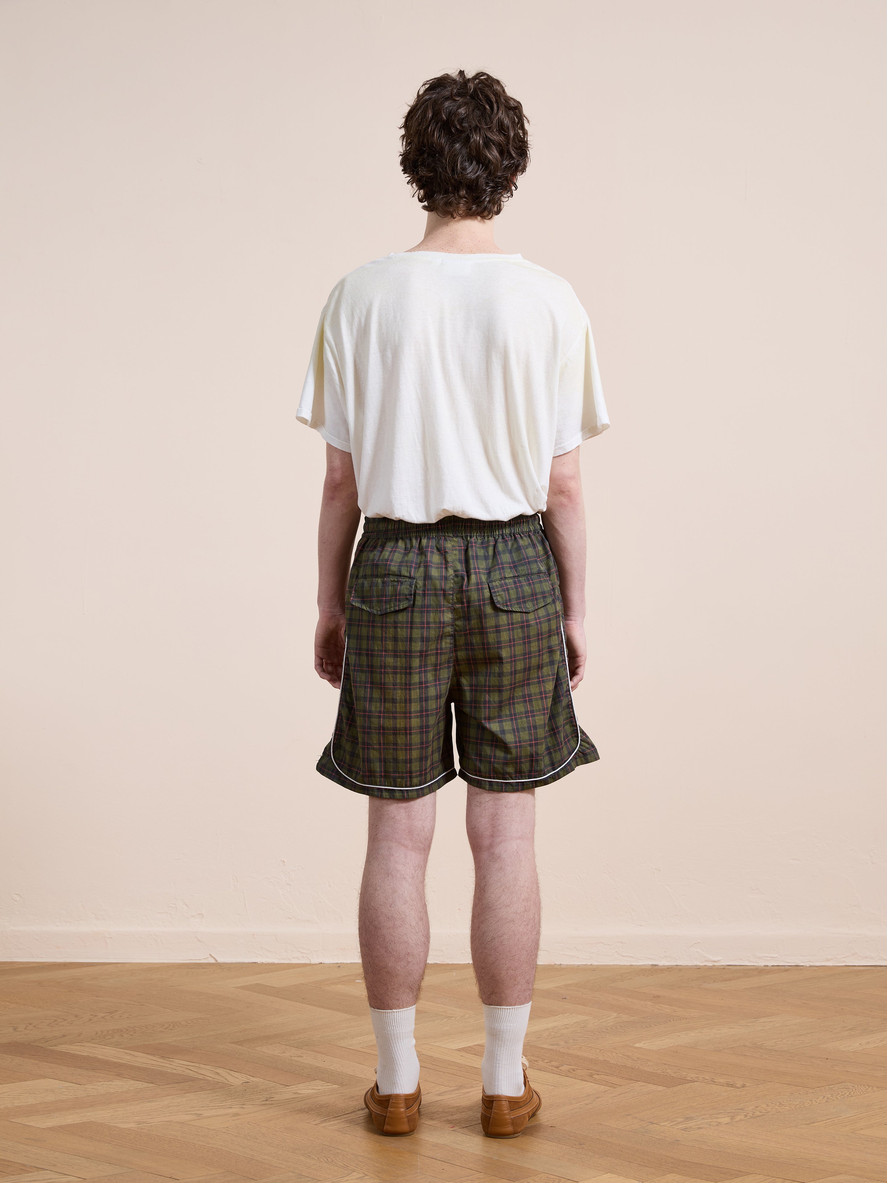 A person stands indoors with their back to the camera, wearing a white t-shirt, white socks, brown shoes, and Found Forest Plaid Track Shorts on a wooden floor against a plain beige wall.