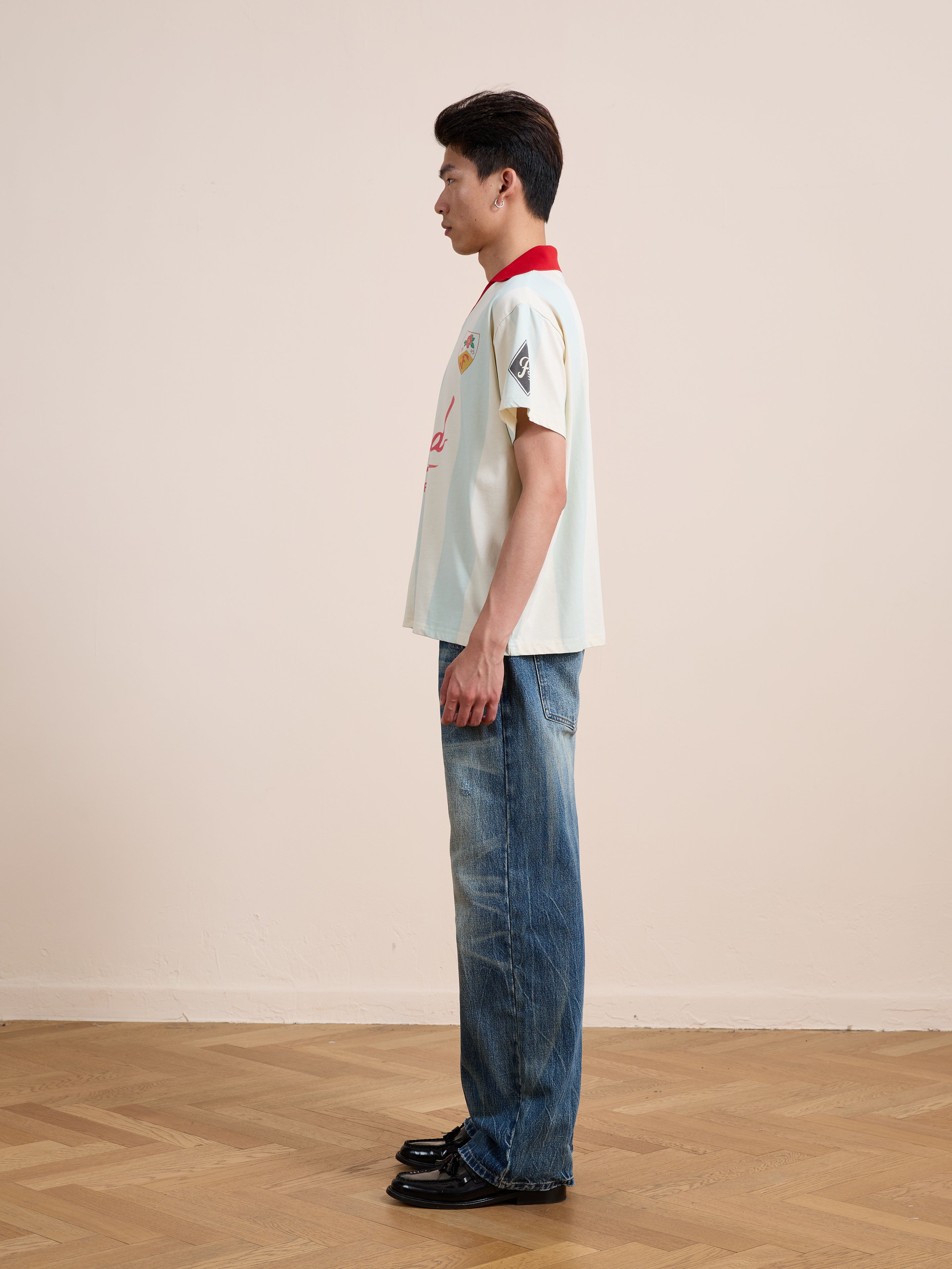 A person standing in profile against a plain wall, wearing the FOUND Found Club Short Sleeve Jersey in cream with a red collar, paired with blue jeans and black shoes on a wooden floor.