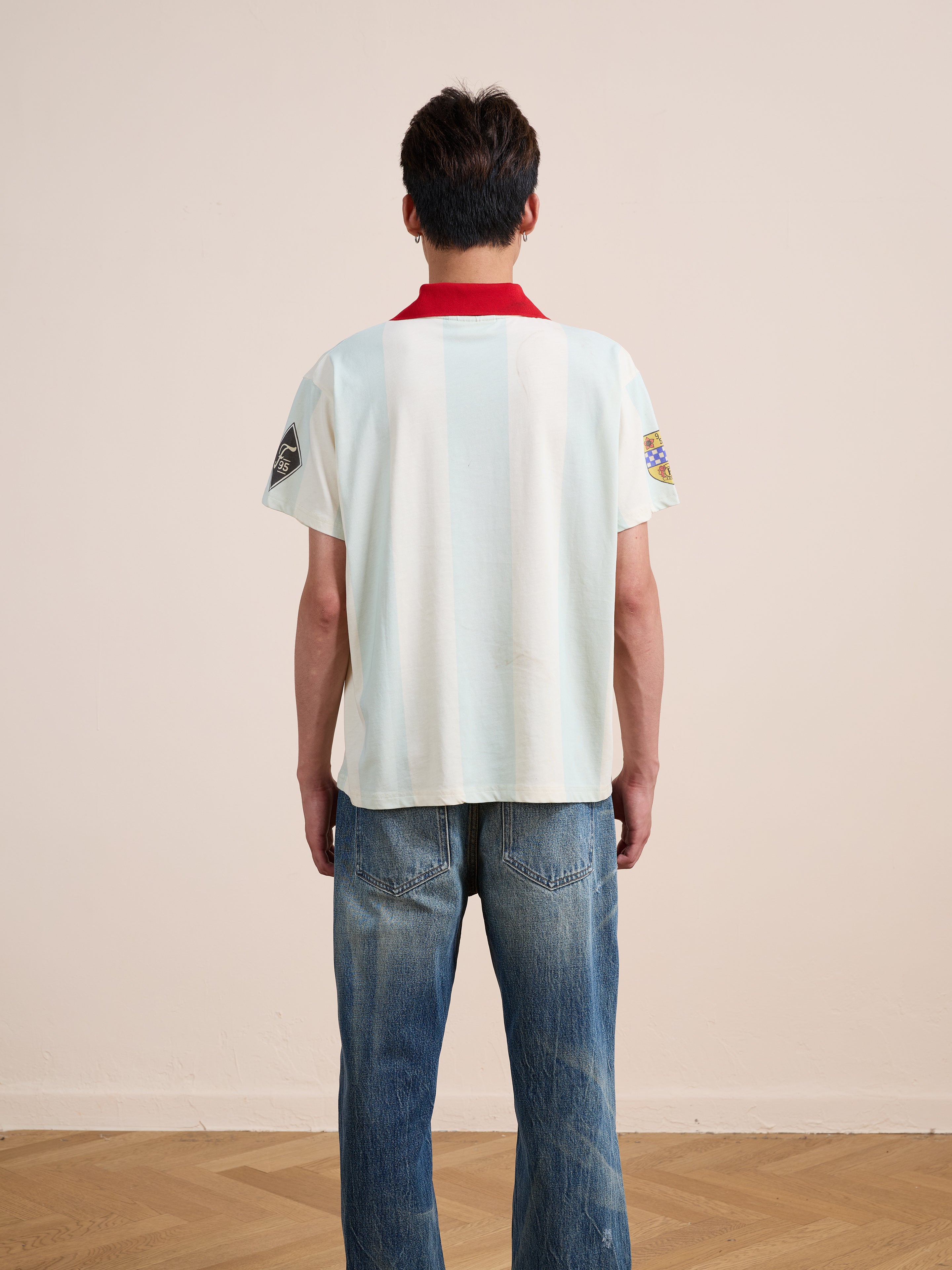 A person stands indoors facing away, wearing the FOUND Found Club Short Sleeve Jersey with a red collar and blue jeans.