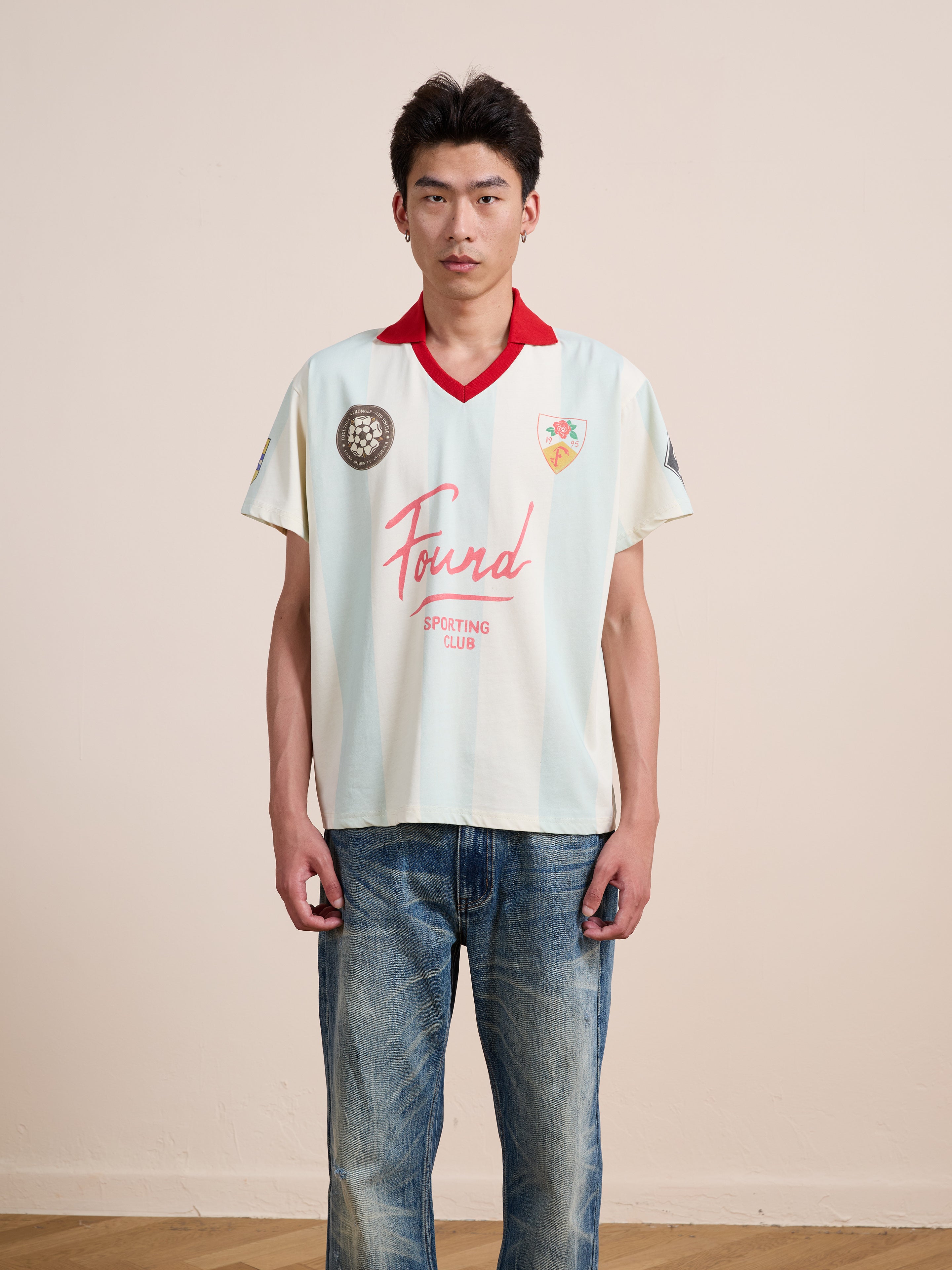 A person stands indoors against a plain wall, wearing the cream-colored FOUND Found Club Short Sleeve Jersey with jeans, facing the camera with a neutral expression. The unisex Found Club Short Sleeve Jersey by FOUND lends their outfit a classic sportswear vibe.
