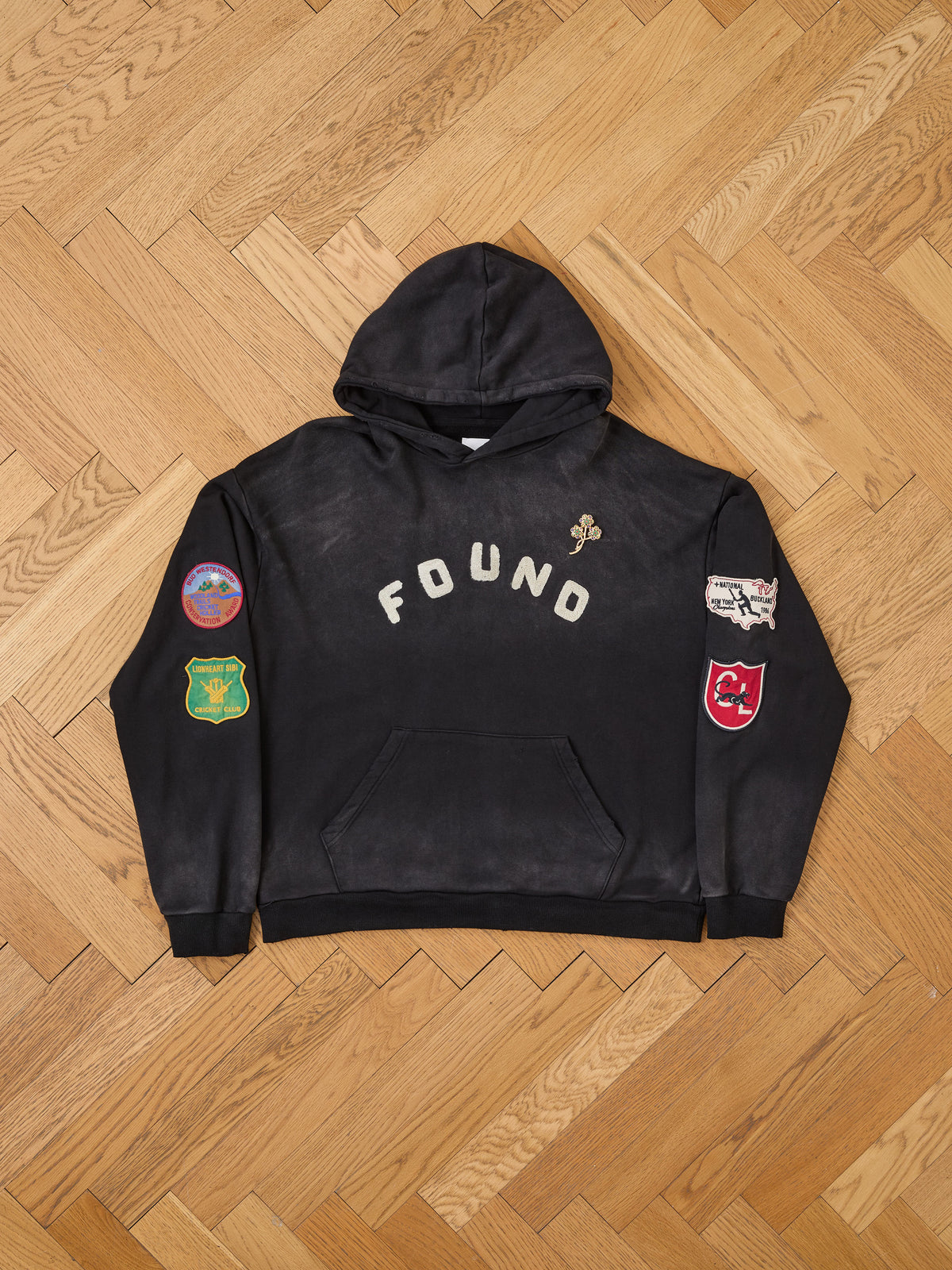 The FOUND Logo Patch Hoodie by FOUND is a unisex, relaxed fit sweatshirt crafted from washed cotton. It features “FOUND” text on the front and four vibrant patches on the sleeves, shown here laid flat on a wooden floor—a versatile everyday staple for any wardrobe.