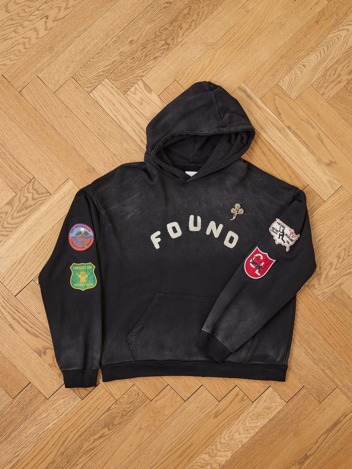 A FOUND Found Logo Patch Hoodie in black, unisex relaxed fit, featuring “FOUND” lettering on the chest and several colorful patches adorning both sleeves, displayed flat on a wooden floor.