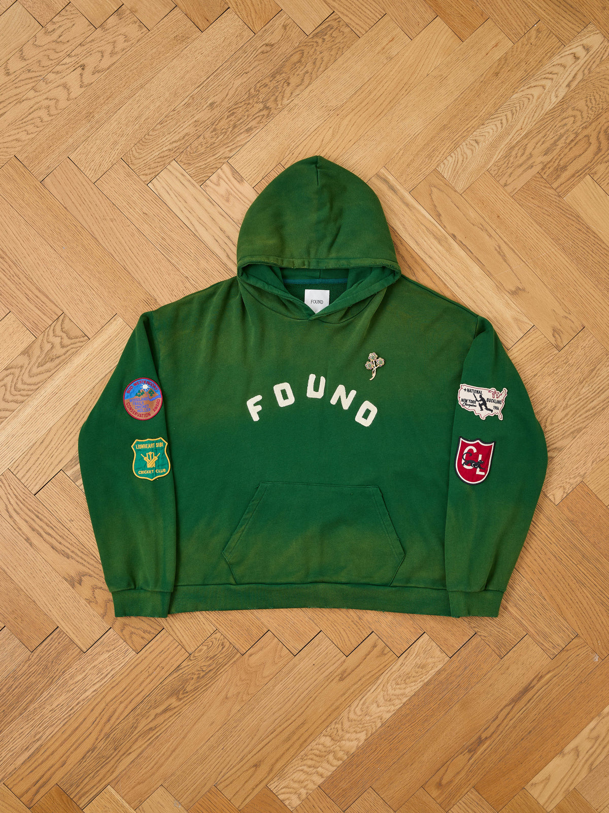 The Found Logo Patch Hoodie by FOUND features a forest green color, “FOUND” text, colorful sleeve patches, and a unisex relaxed fit.