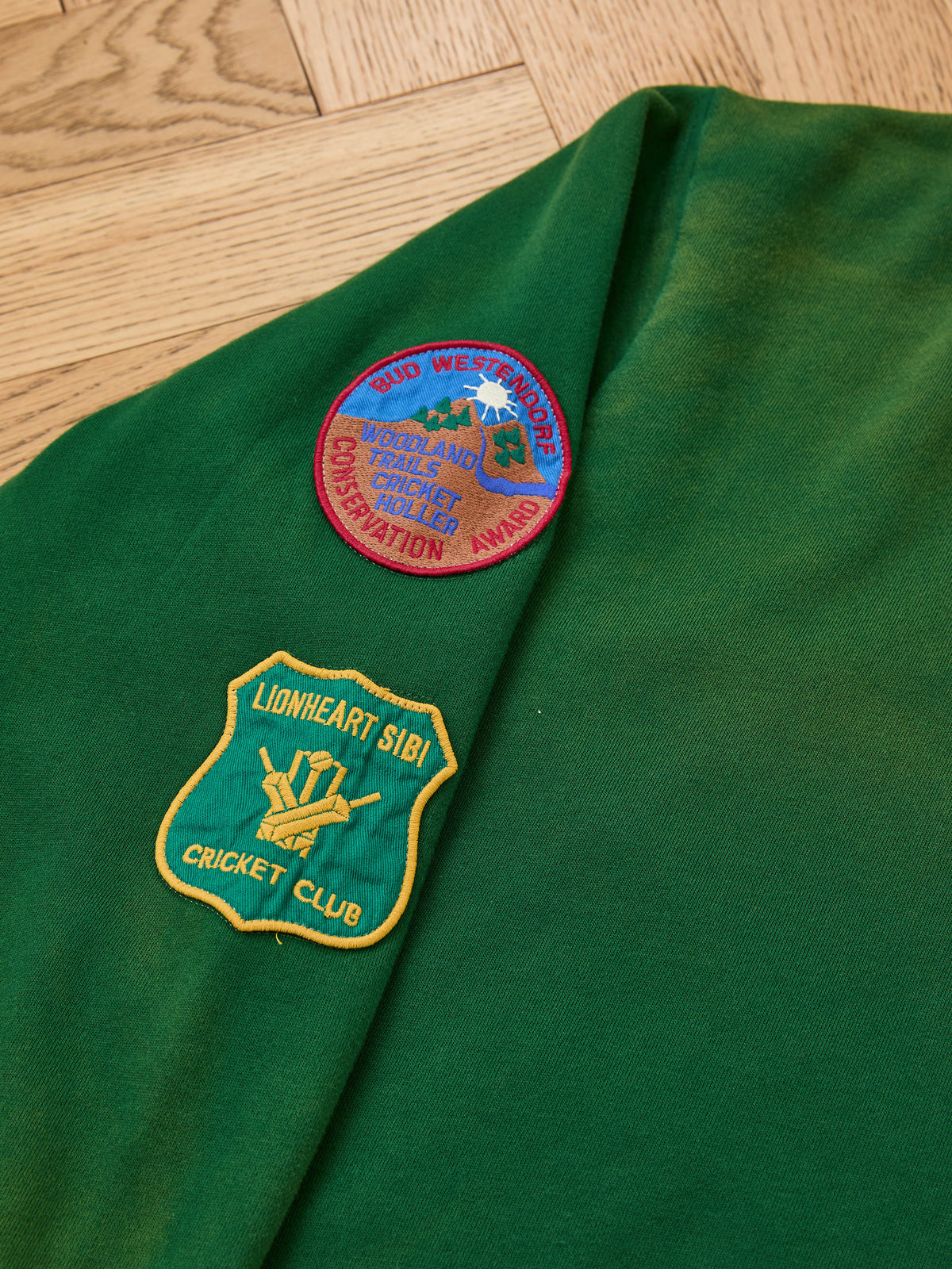 The Found Logo Patch Hoodie by FOUND in vintage forest green, features two sewn patches—a conservation award and Lionheart Sibi Cricket Club—on a wooden surface.