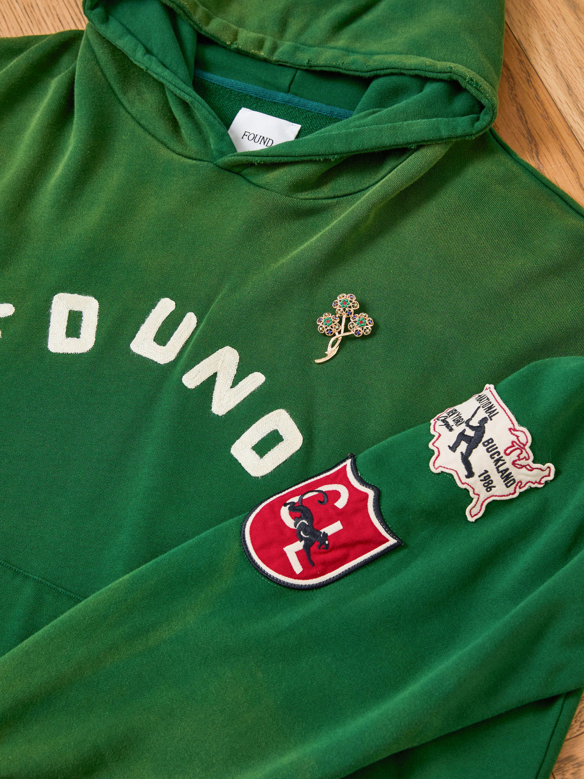 FOUND Found Logo Patch Hoodie in vintage forest green, featuring a relaxed unisex fit, “FOUND” text, clover pin, red shield horse patch, and a white USA-shaped bull graphic.