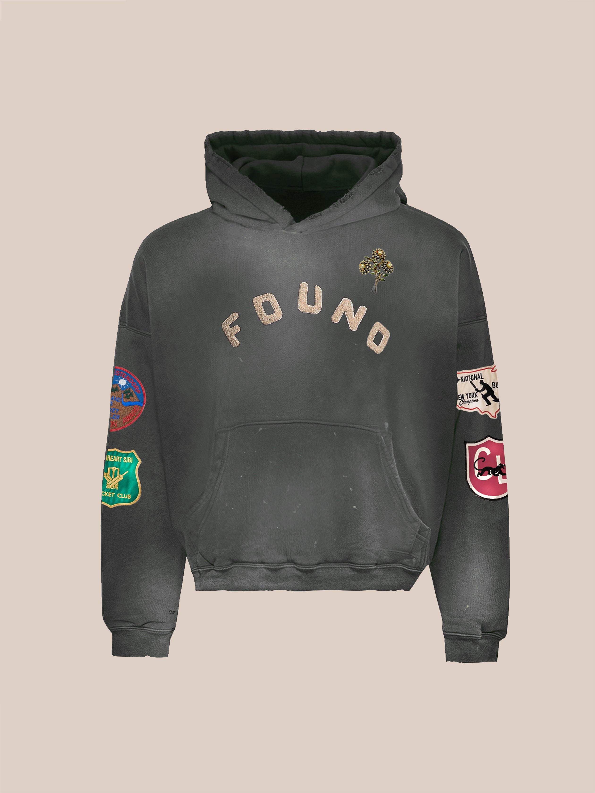 The FOUND Found Logo Patch Hoodie is a faded black unisex hoodie with "FOUND" on the chest, colorful sleeve patches, and a front kangaroo pocket. This relaxed fit washed cotton piece is shown against a plain beige background.