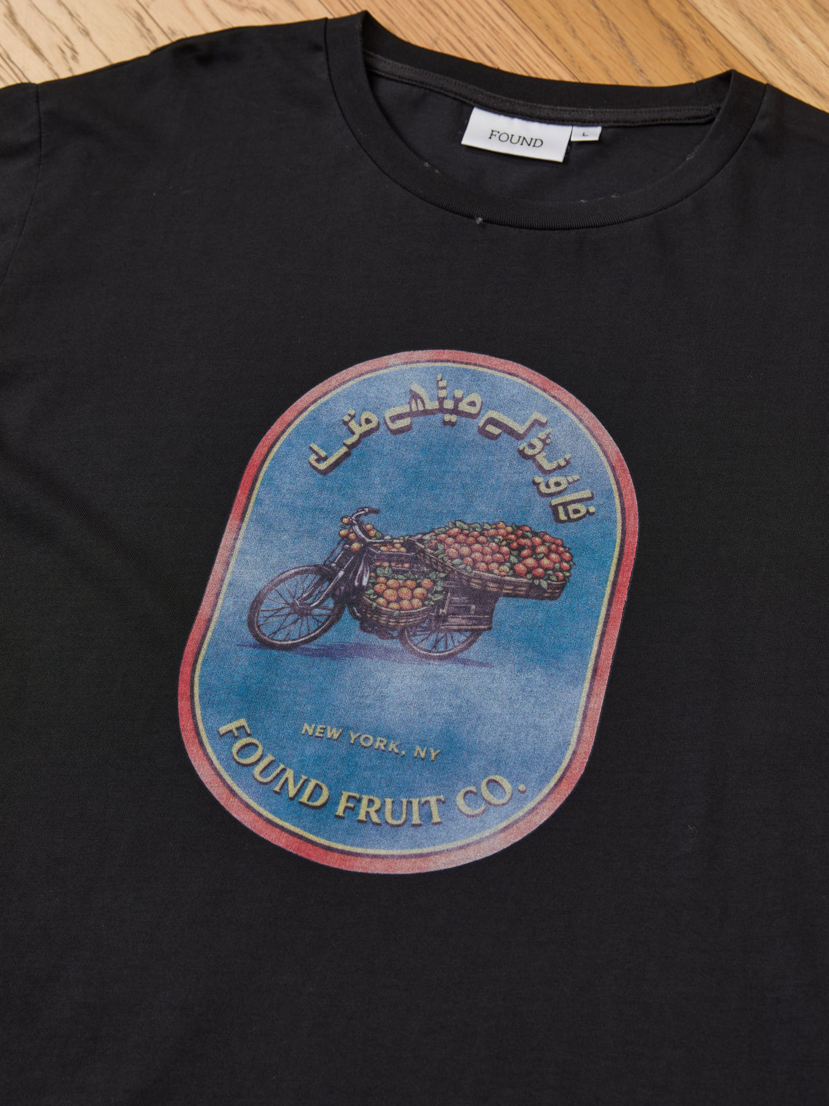 The Fruit Co Tee by FOUND is a unisex relaxed fit shirt, featuring a bicycle with fruit crates and “FOUND FRUIT CO., New York, NY” in English and Arabic.