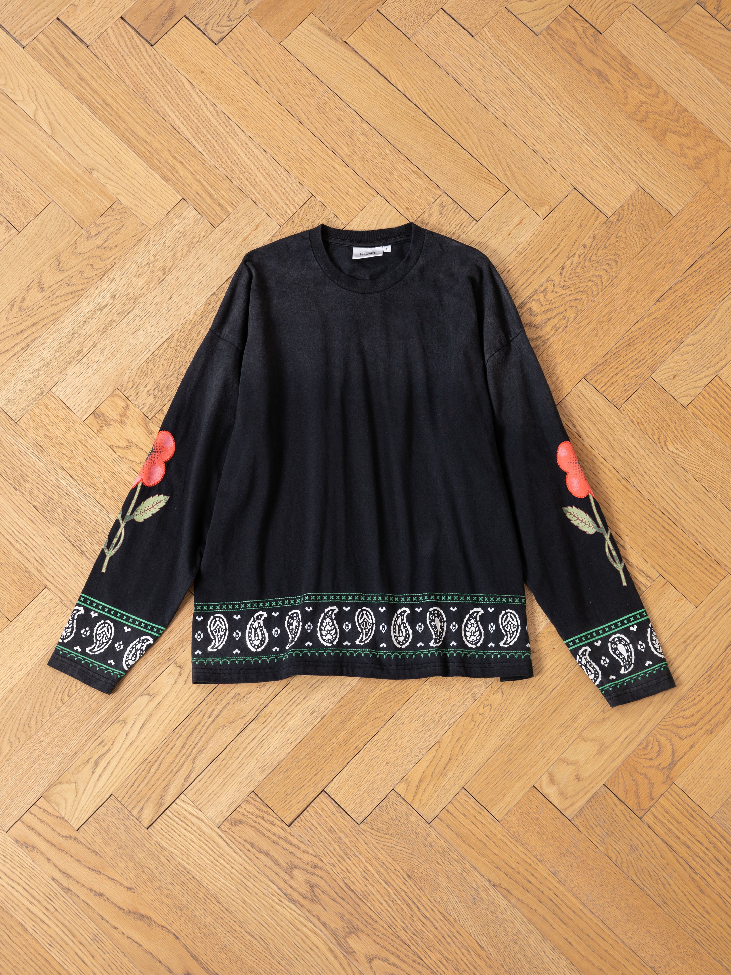 The FOUND Garden Bloom Embroidered LS Tee features floral embroidery on the sleeves, a white paisley pattern with green trim at the hem and cuffs, and a relaxed fit, shown laid flat on a wooden floor.