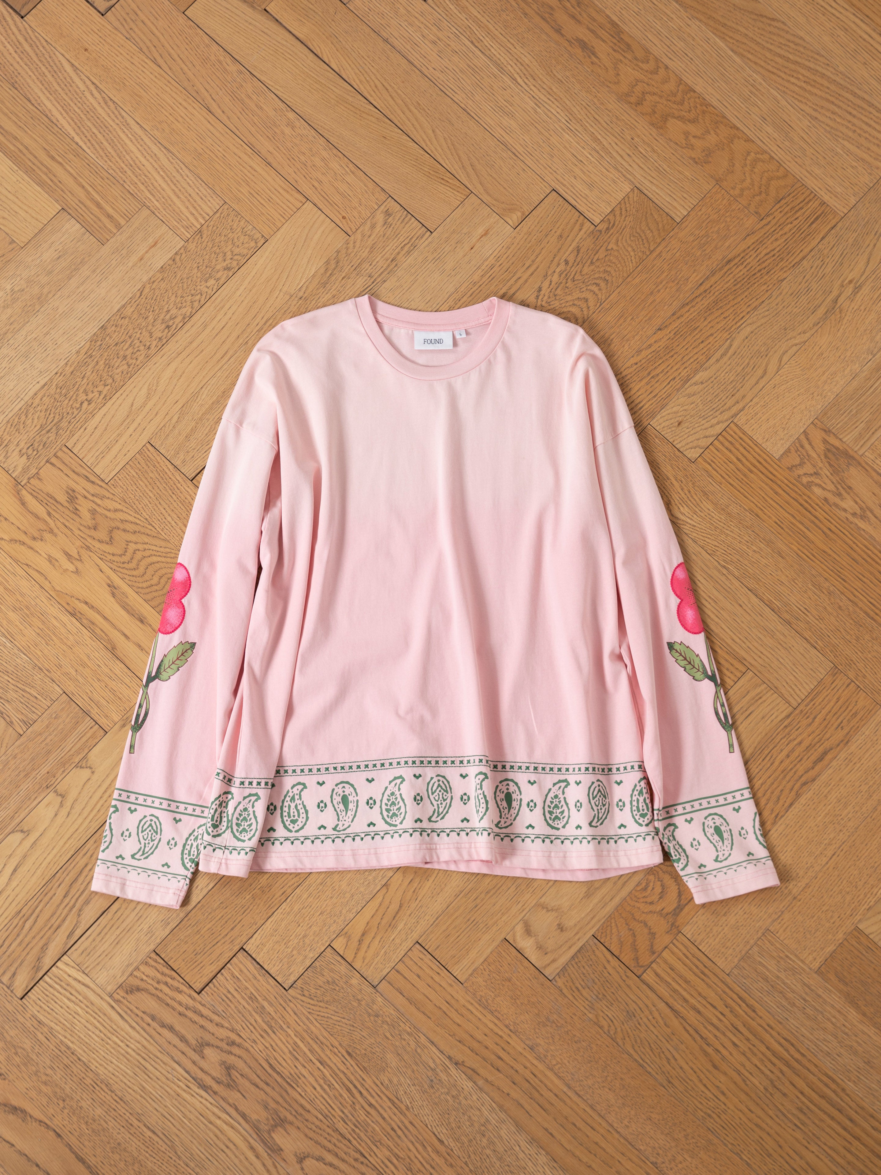 The FOUND Garden Bloom Embroidered LS Tee in light pink features floral sleeve graphics and a paisley hem border, shown laid flat on a wooden herringbone floor in a unisex relaxed fit.