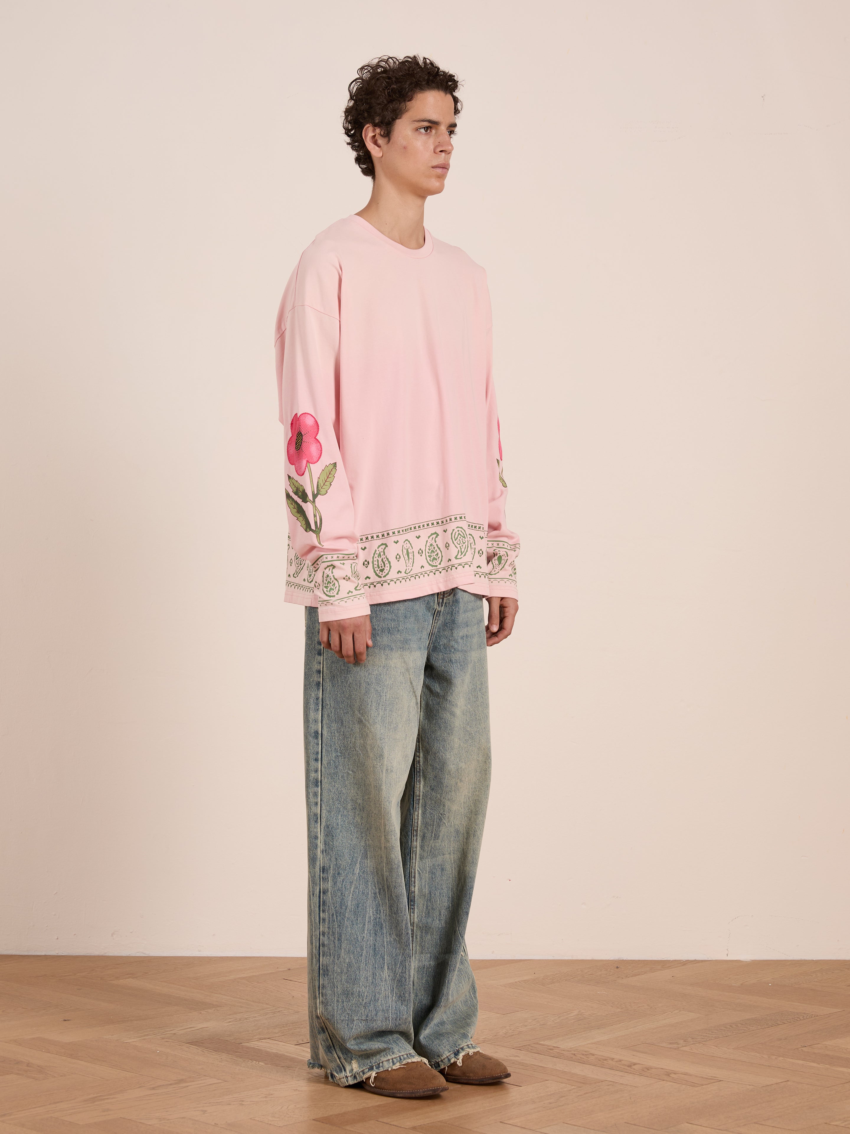 Indoors, a person wears the FOUND Garden Bloom Embroidered LS Tee—a unisex relaxed fit top with floral embroidery and lace trim—paired with wide-leg faded jeans and brown shoes.
