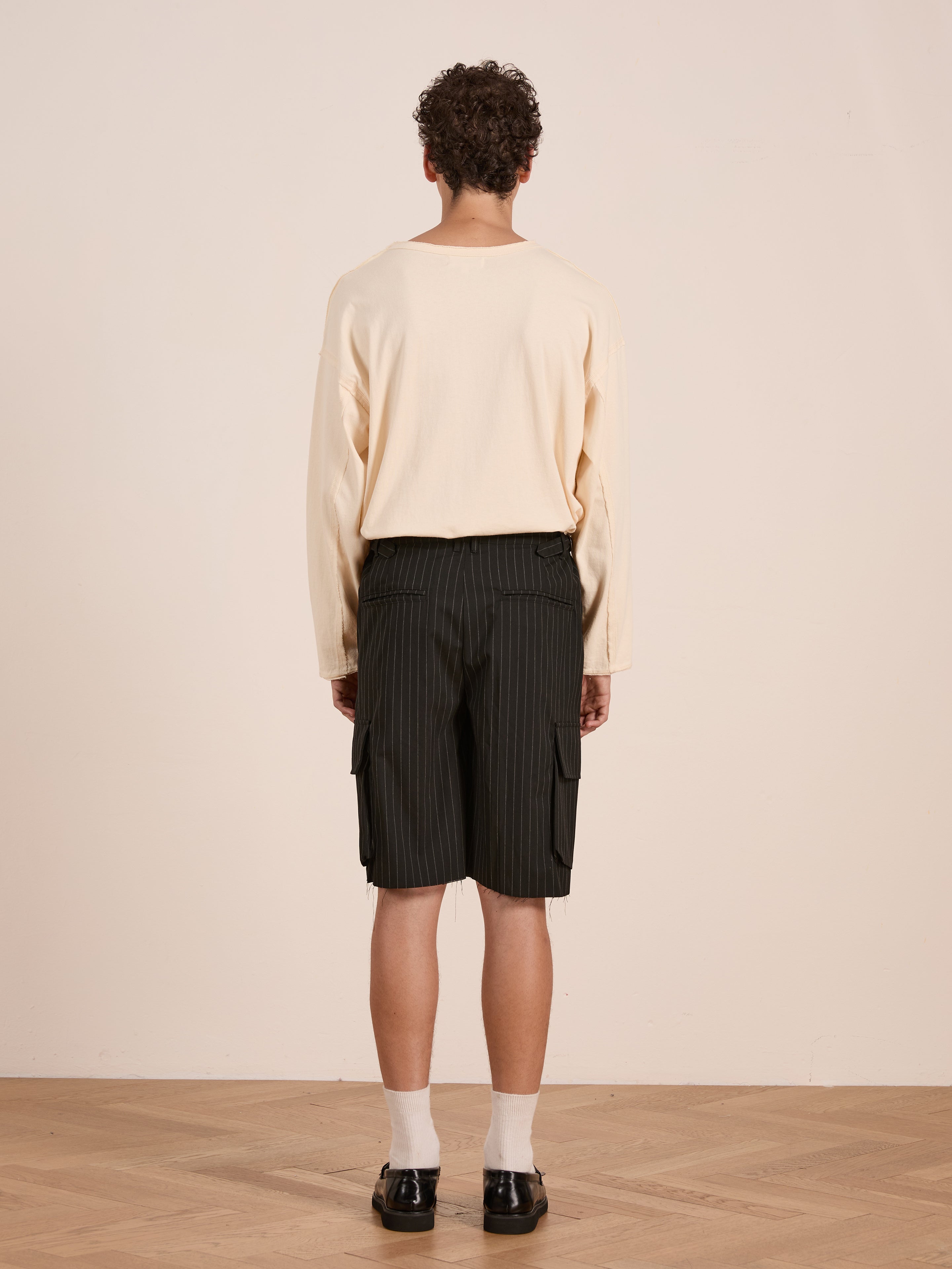A person stands indoors facing away, wearing a cream long-sleeve shirt, FOUND Hava Pinstripe Pleated Cargo Trouser Shorts with an adjustable fit, white socks, and black shoes.
