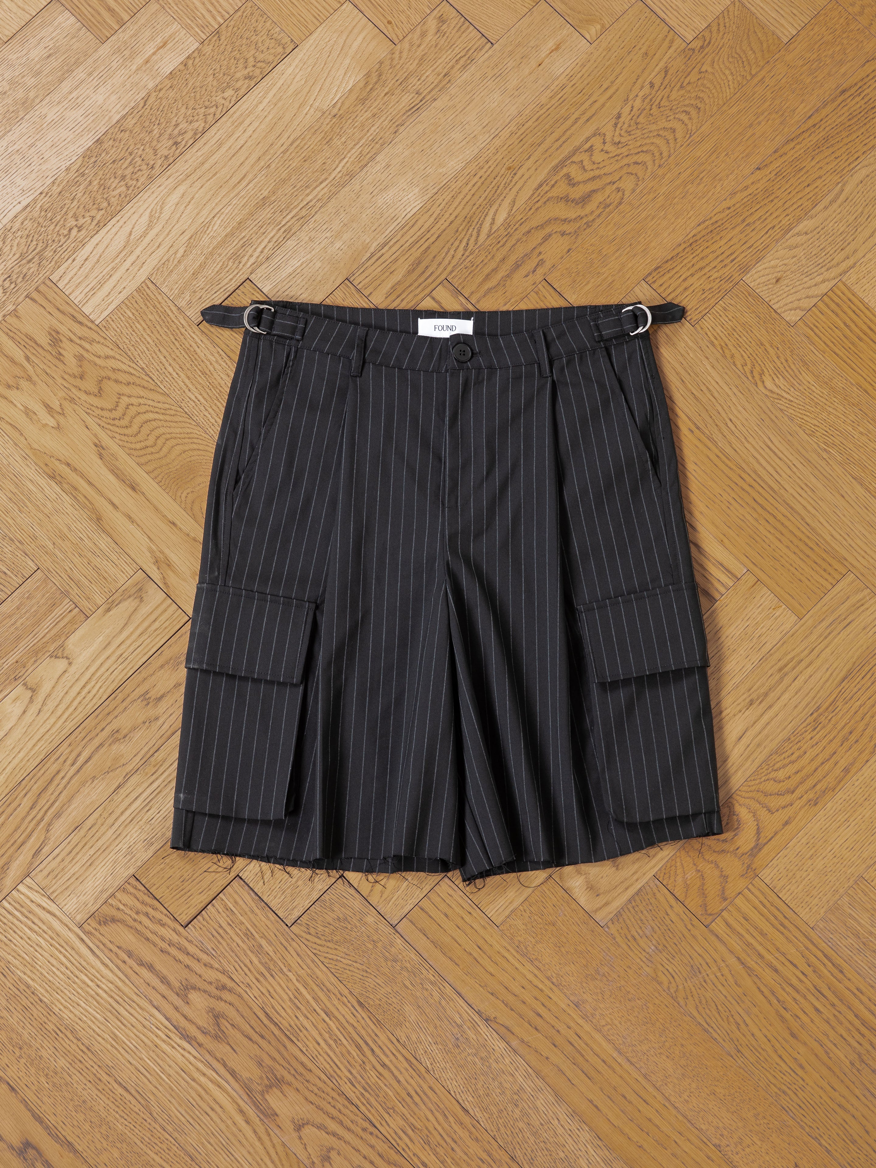 FOUND's Hava Pinstripe Pleated Cargo Trouser Shorts, featuring flap pockets and belt loops, are unisex shorts with an adjustable fit—combining classic style and modern utility for versatile wear. Shown laid flat on a herringbone wood floor.